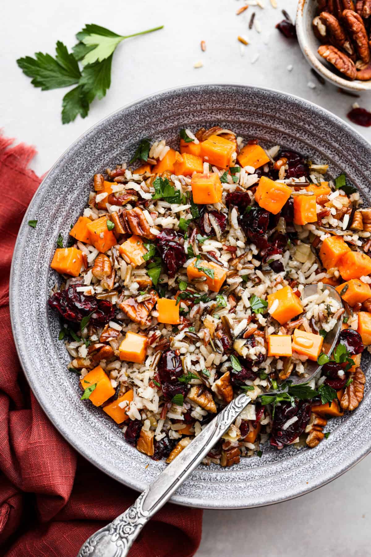 Cranberry pecan sweet potato wild rice pilaf is a perfect fall side dish infused with incredible flavors and textures. Crunchy pecans, cranberries, sweet potato, wild rice, and herbs come together in this tasty dish! Top view of cranberry pecan sweet potato wild rice pilaf in a gray bowl with a in the bowl spoon.