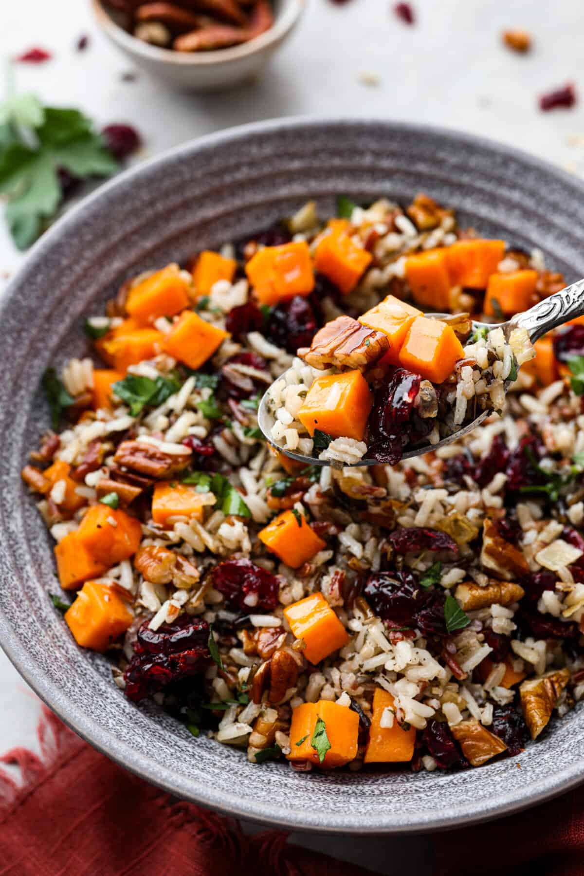 Cranberry pecan sweet potato wild rice pilaf is a perfect fall side dish infused with incredible flavors and textures. Crunchy pecans, cranberries, sweet potato, wild rice, and herbs come together in this tasty dish! Side view of cranberry pecan sweet potato wild rice pilaf in a gray bowl with a spoon. The spoon is lifting up a bite of pilaf.