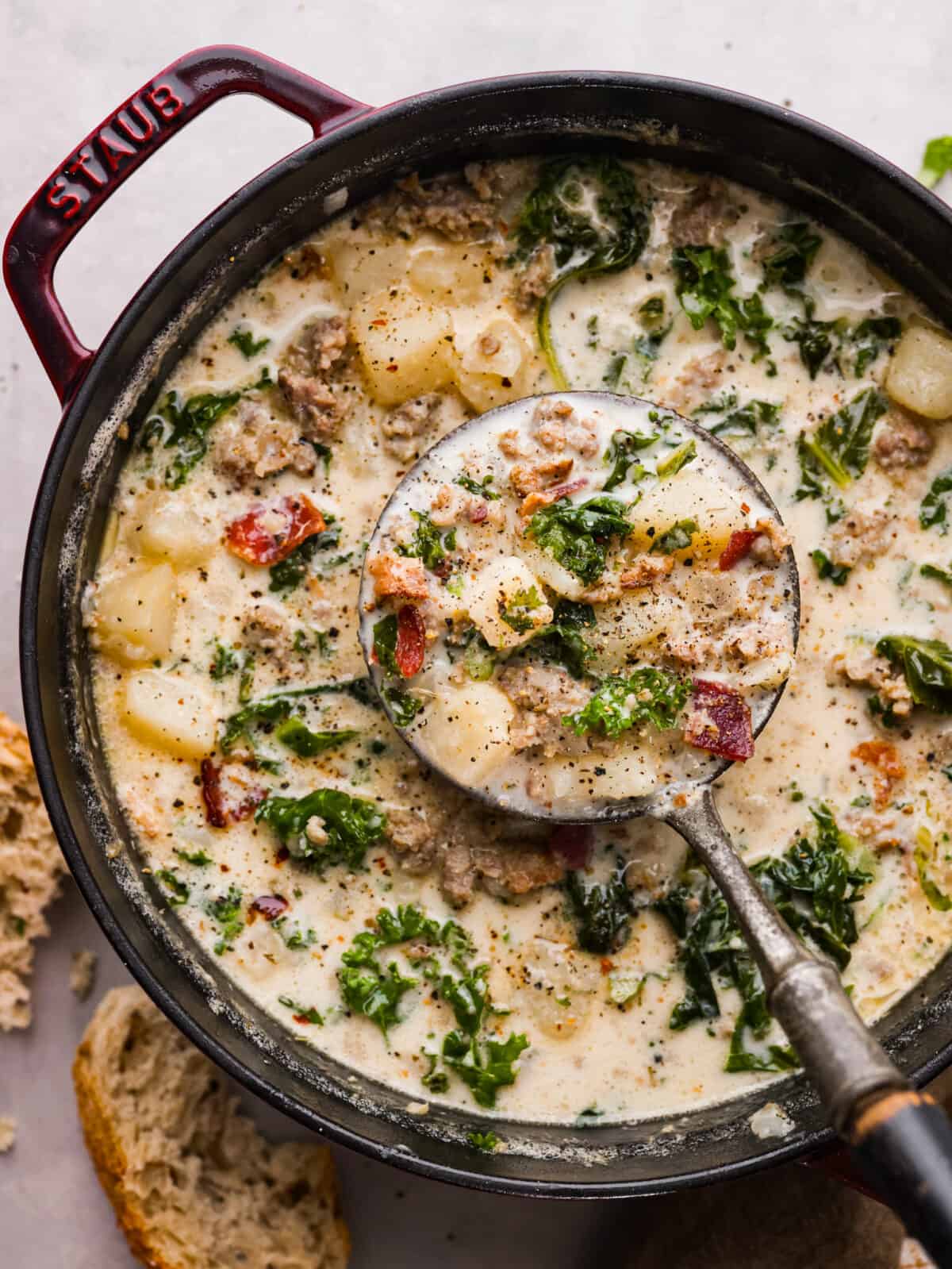 Creamy Zuppa Toscana is a savory Italian soup that is made with potatoes, spicy sausage, bacon, and kale. It is thick, creamy, and delicious and is a must-make soup this fall! The top view of Zuppa Toscana soup in a pot with a ladle scooping some out.