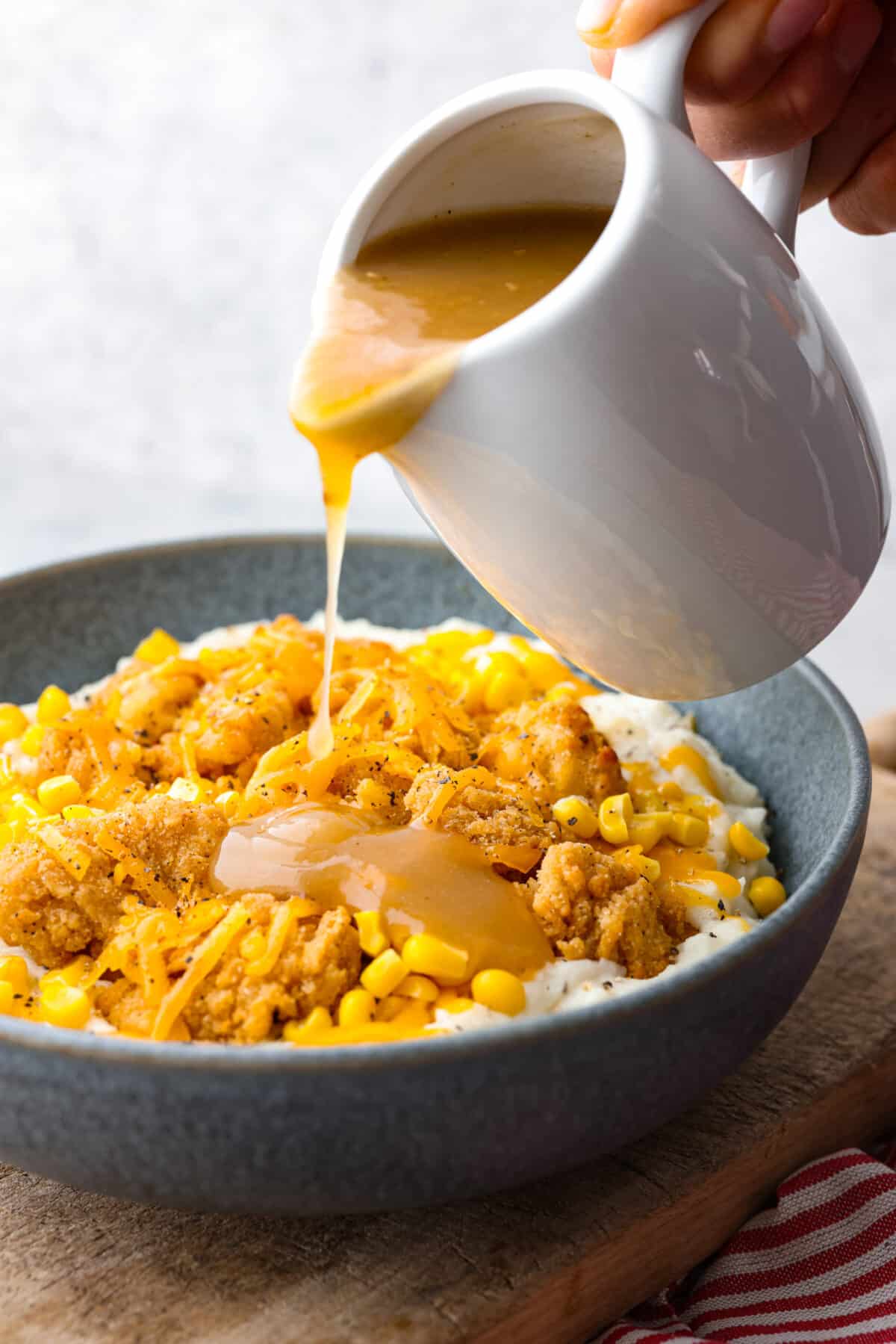 This irresistible copycat KFC Famous Bowl is super quick and easy to make at home and tastes even better than the fast food classic. It's a warm & comforting dinner that the whole family will enjoy! Pouring gravy over chicken, corn, and mashed potatoes.