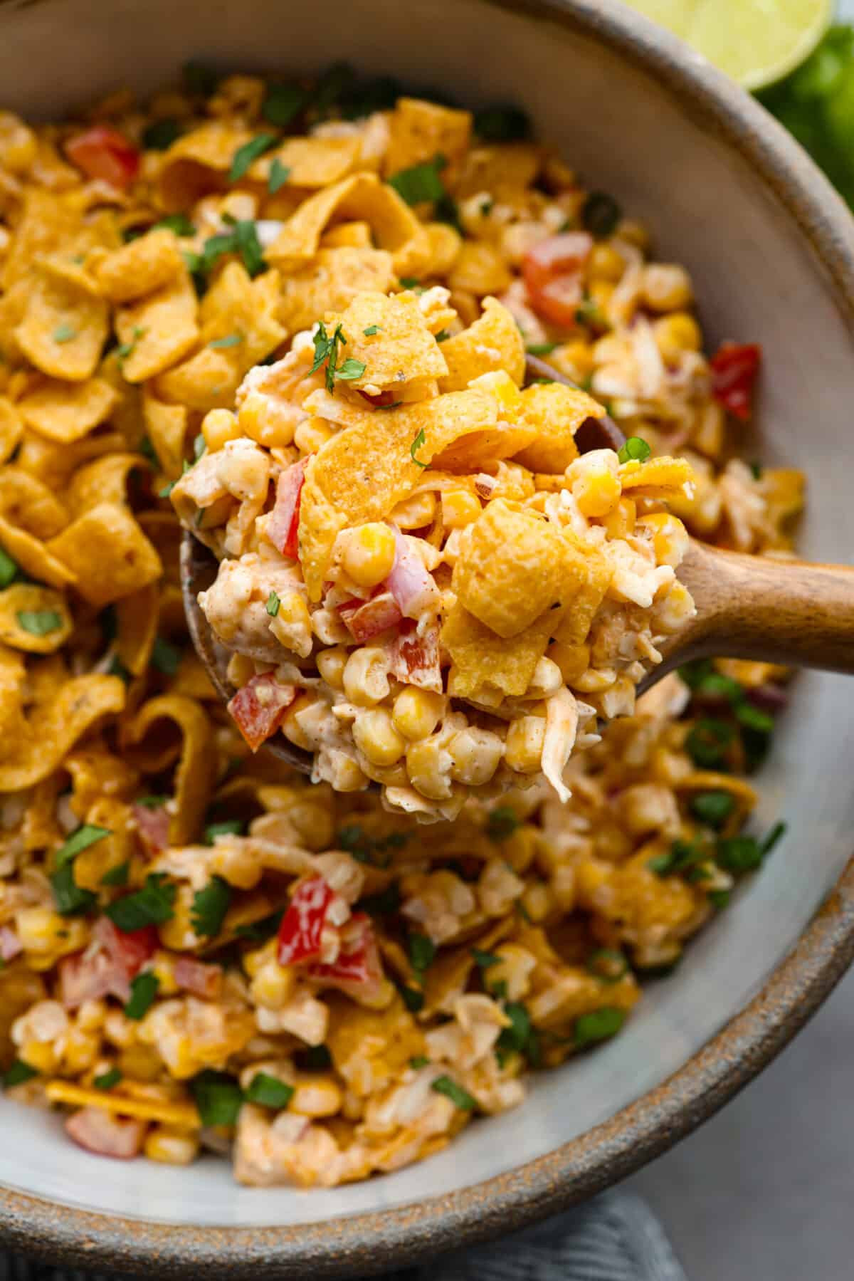 Crunchy Frito corn salad is a fun side dish bursting with Mexican-inspired flavors! Corn, cheese, peppers, and onion get tossed in a tasty dressing and mixed with Fritos corn chips. A surprising FAVORITE at any get-together! Close view of frito corn salad in a bowl. A wood spoon is lifting up a scoop of salad.