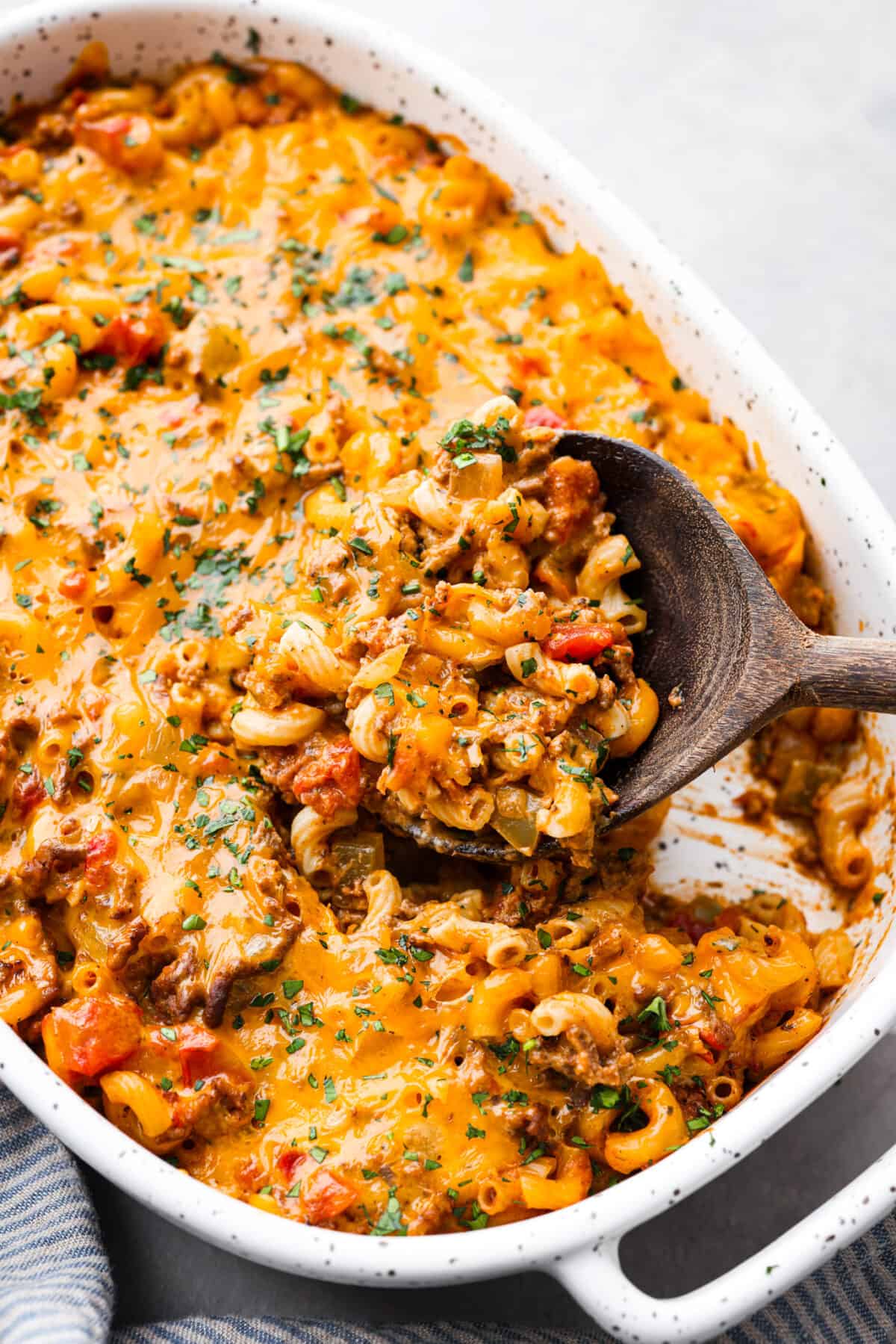 Cheeseburger Casserole has all of those delicious cheeseburger flavors in one easy-to-make dish! Loaded with ground beef, veggies, dill relish, pasta and plenty of cheese, all baked together for a comforting family-friendly dinner. Taking a scoop of cheeseburger casserole with a wooden spoon.