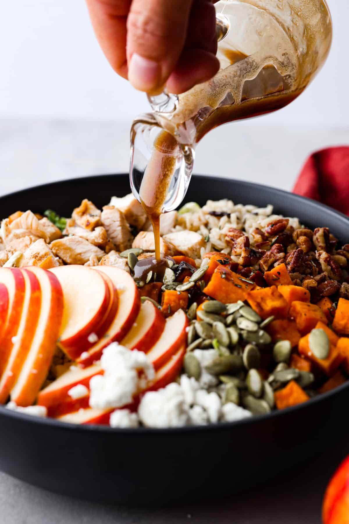 This gorgeous Harvest Bowl has all the most delicious flavors of fall packed in one easy salad. It’s filled with creamy sweet potatoes, kale, Brussels sprouts, wild rice, grilled chicken, sweet apple, cheese, crunchy candied pecans, and pepitas with the best balsamic vinaigrette dressing. You'll be craving it all season long! Pouring dressing over a harvest bowl.