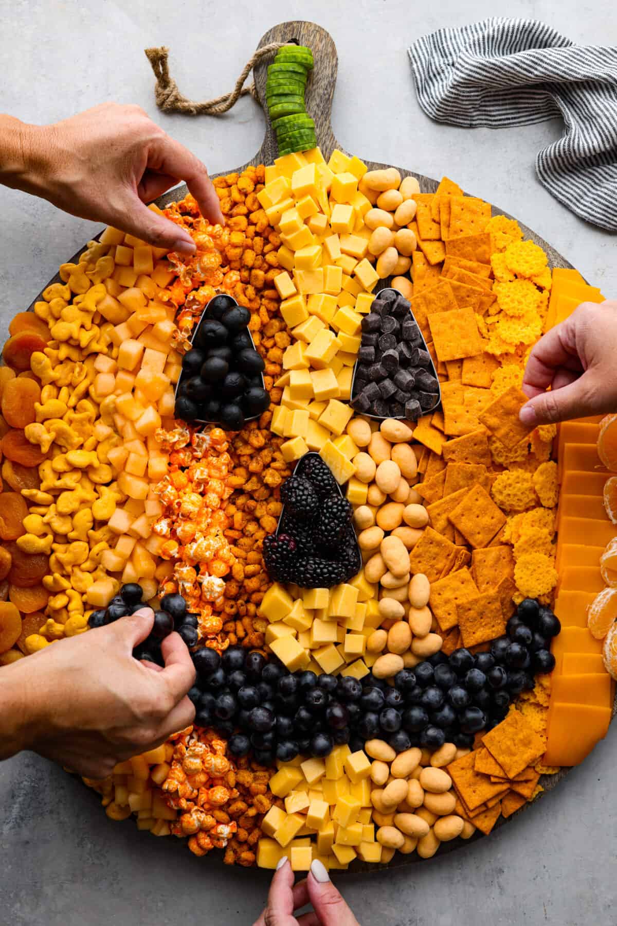 Halloween is the BEST time to get creative and have fun with your food! Gather all things orange and bring this pumpkin charcuterie board to your next fall get-together. Fill your board to look like a Jack-o-lantern; it will be the talk of the party! Overhead view of pumpkin charcuterie board with four hands reaching into grab snacks.