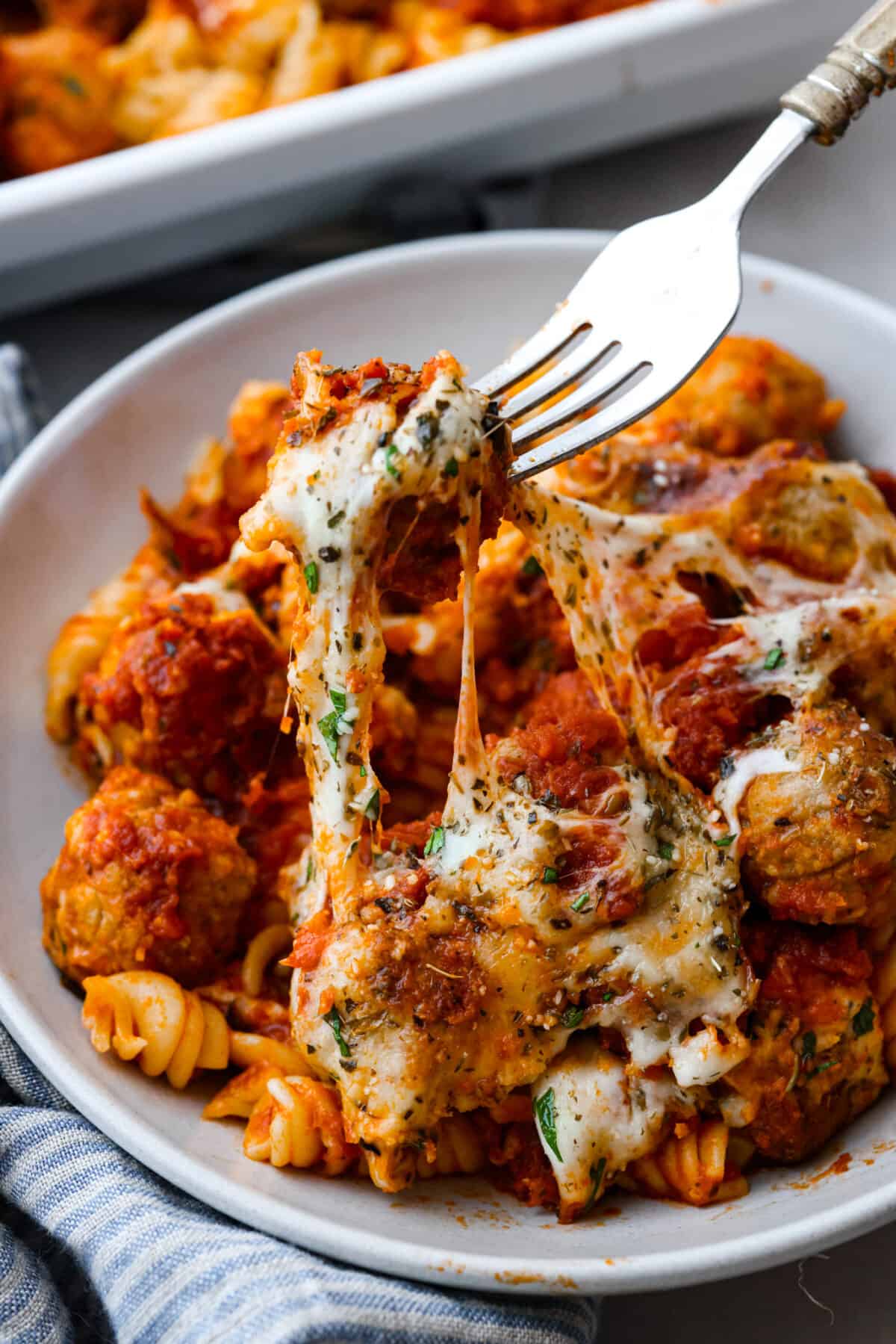With just one dish to wash and a handful of simple ingredients, you can treat your entire family to this hearty meatball casserole. It has tender meatballs nestled in a bed of rotini pasta, smothered in marinara sauce, and topped with gooey mozzarella and parmesan cheese. Picking up a cheesy meatball with a fork.