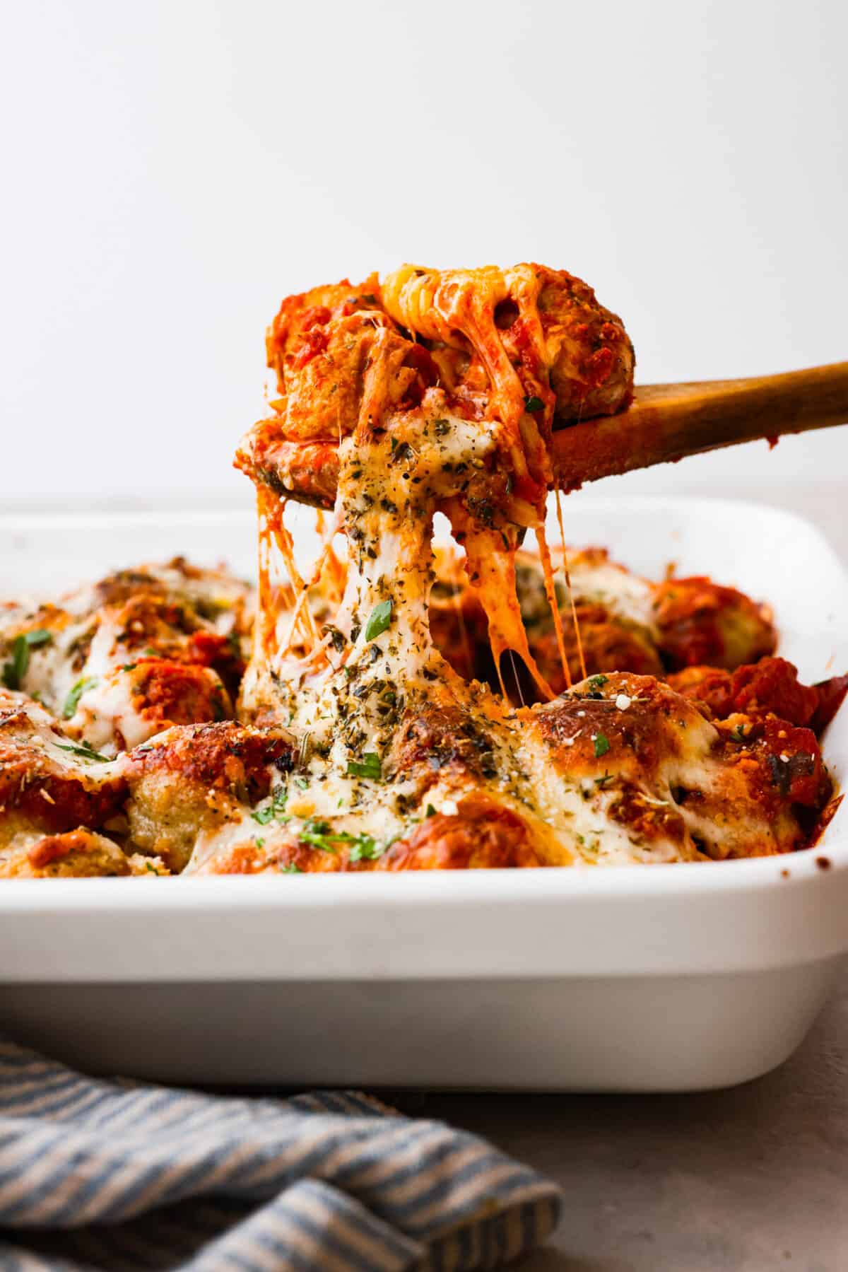 With just one dish to wash and a handful of simple ingredients, you can treat your entire family to this hearty meatball casserole. It has tender meatballs nestled in a bed of rotini pasta, smothered in marinara sauce, and topped with gooey mozzarella and parmesan cheese. Scooping up meatballs and cheese with a wooden spoon.