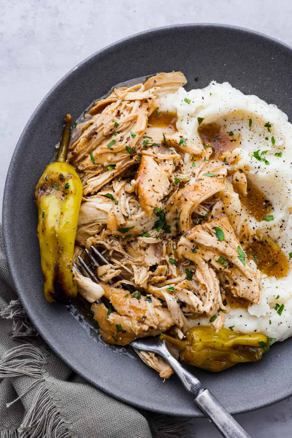 Crockpot Mississippi Chicken is all about fork-tender, juicy chicken that's bursting with flavor. You've got zesty pepperoncini peppers, ranch seasoning, au jus mix, and butter that slow cooks with it for some seriously amazing flavor. It's the perfect Southern comfort meal! Shredded chicken served on a gray plate with mashed potatoes.