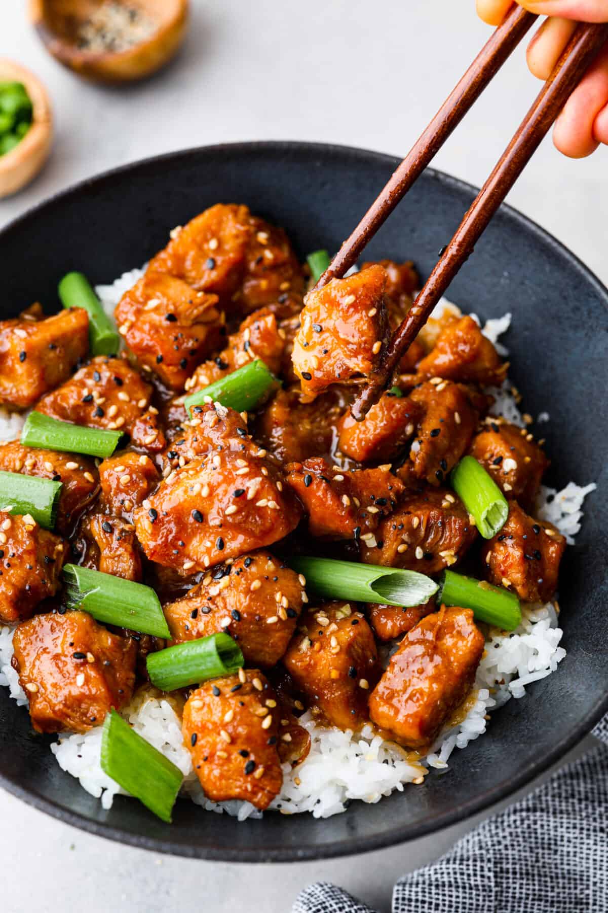 Slow cooker General Tso's chicken is a super easy version of everyone's favorite Chinese entree! Crispy chicken cooked in a sweet and savory sauce with a little added heat. This is way better than takeout! Close view of slow cooker General Tso's chicken in a black bowl served over rice. Garnished with sesame seeds and green onions. Chopsticks are picking up a piece of chicken.