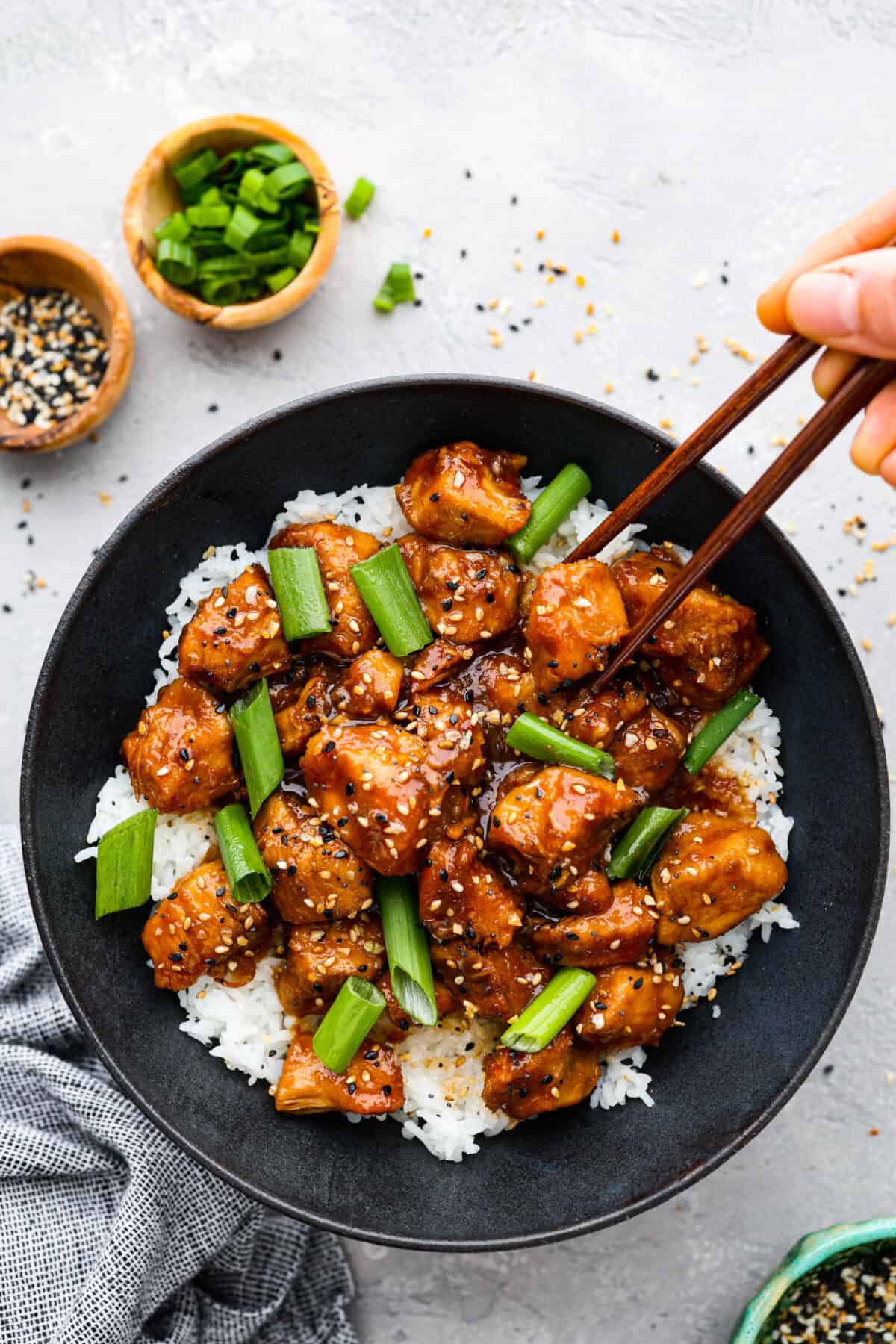 Slow cooker General Tso's chicken is a super easy version of everyone's favorite Chinese entree! Crispy chicken cooked in a sweet and savory sauce with a little added heat. This is way better than takeout! Top view of slow cooker General Tso's chicken in a black bowl served over rice. Garnished with sesame seeds and green onions. Chopsticks are picking up a piece of chicken.