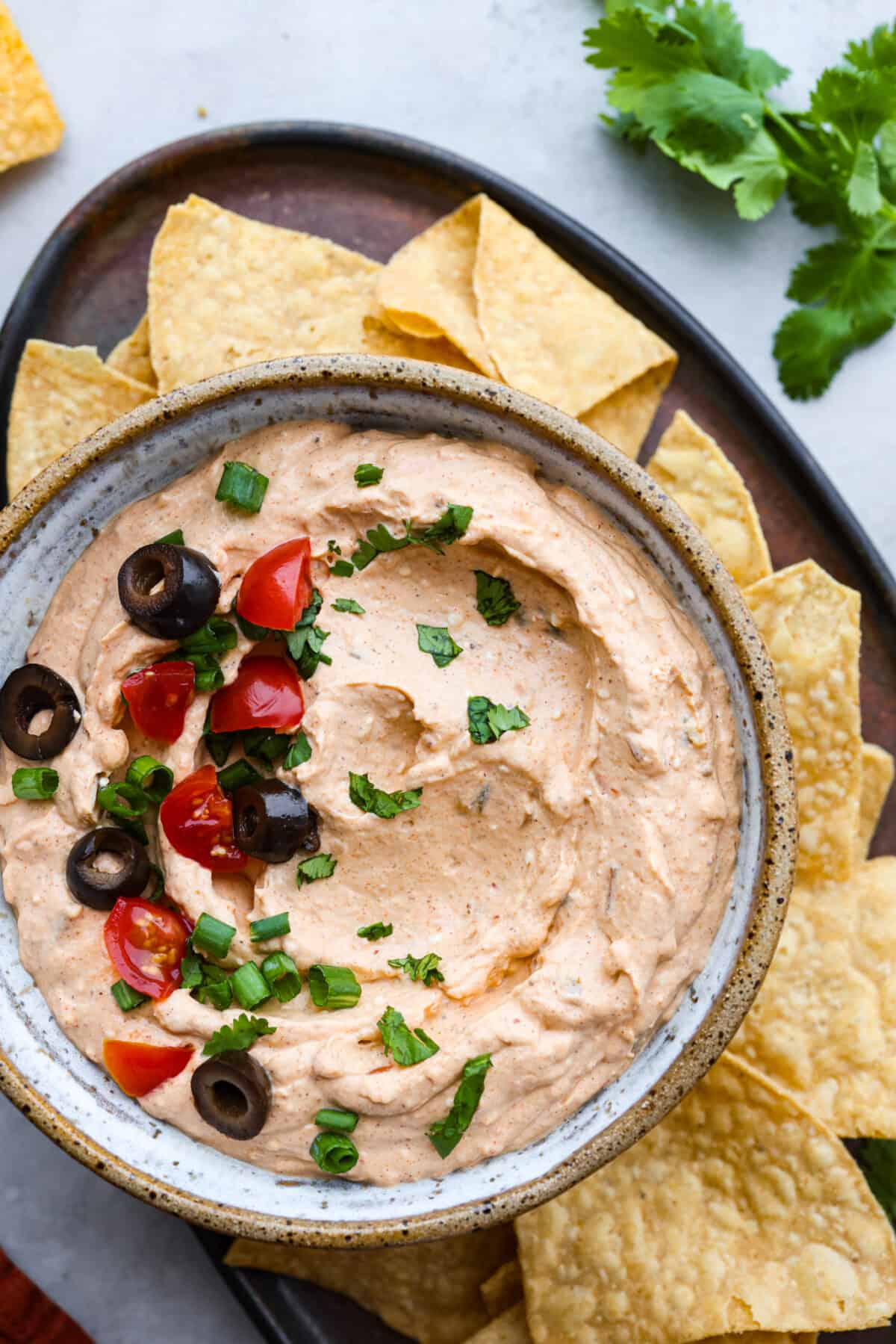 Taco dip takes all the tasty flavor of tacos and turns it into a creamy, zesty, easy-to-eat appetizer. Whip this up in 5 minutes with just 4 key ingredients. It is so deliciously addicting and will be the star of the party! Hero image of taco dip garnished with tomatoes, olives, and cilantro in a stoneware bowl. It is surrounded by tortilla chips.