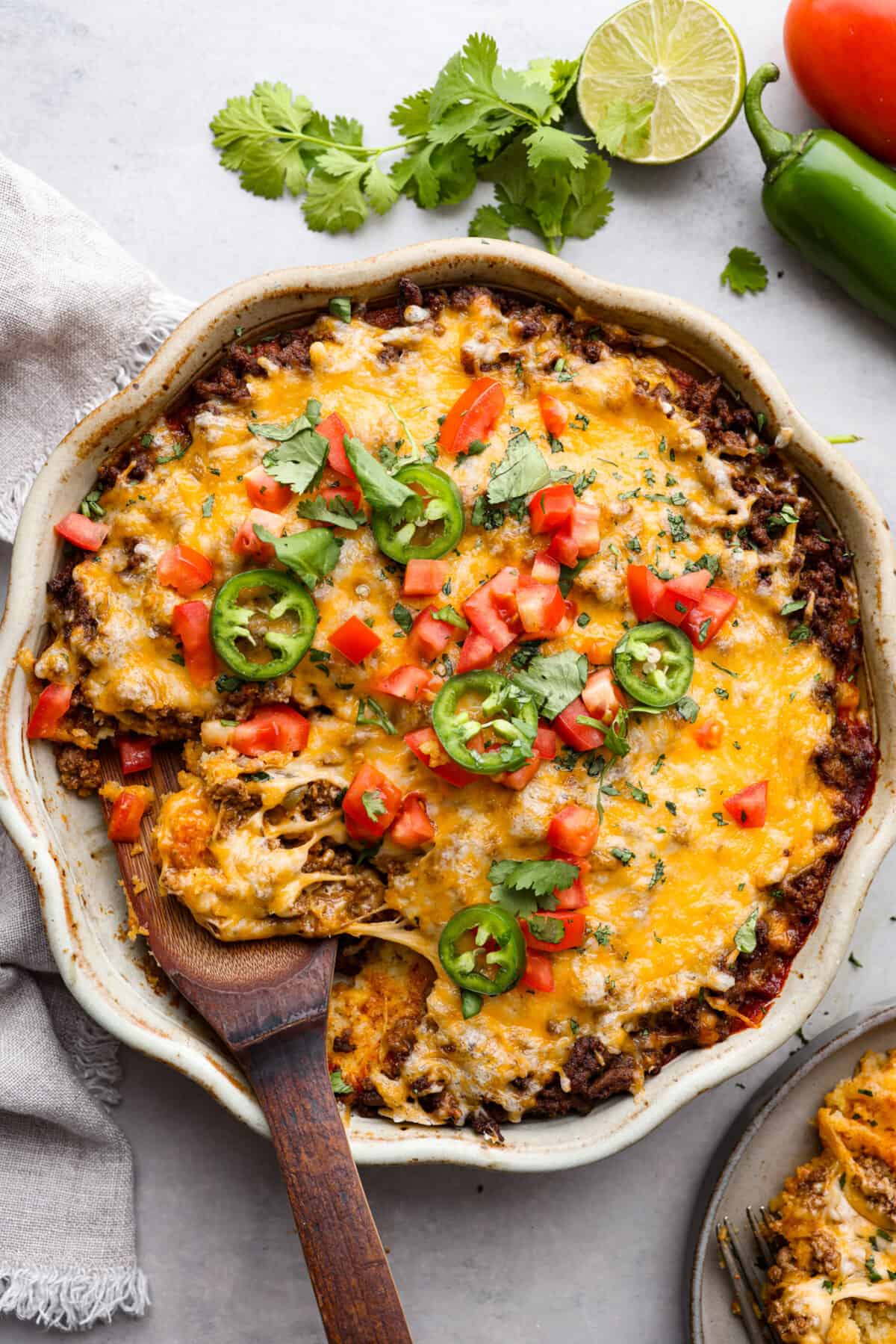 Tamale Pie is seasoned ground beef on top of a cornbread crust, drenched in a bold enchilada sauce, and topped with cheese and tomatoes. This casserole recipe is an easy spin on tamales, making it perfect for a weeknight dinner. The top view of tamale pie that is garnished with jalapeรฑos, cilantro and tomatoes.