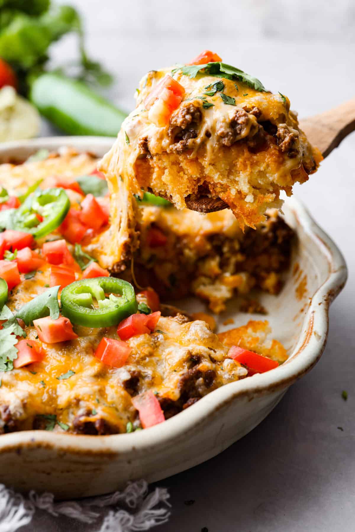 Tamale Pie is seasoned ground beef on top of a cornbread crust, drenched in a bold enchilada sauce, and topped with cheese and tomatoes. This casserole recipe is an easy spin on tamales, making it perfect for a weeknight dinner. Tamale Pie being scooped out of a casserole dish with a wooden spoon.