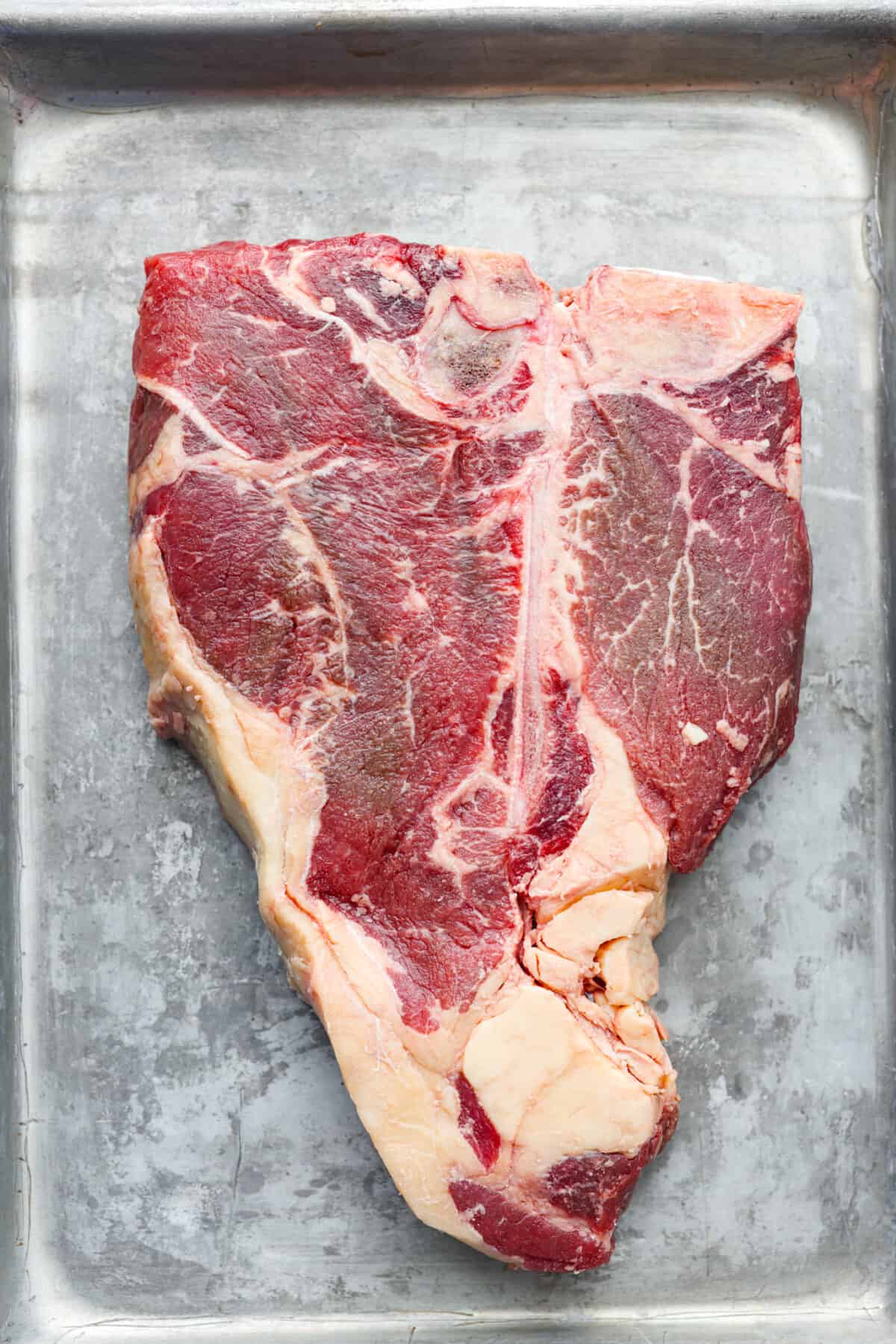 Indulge in the ultimate T-bone steak experience with this recipe! It takes an already awesome cut of meat and cooks it to perfection. The mix of olive oil, spices, and a hot skillet gives you a beautifully seared crust. Top it off with some garlic butter and fresh herbs, then you have the perfect meal ready for any occasion! Top-down view of a raw tbone steak.