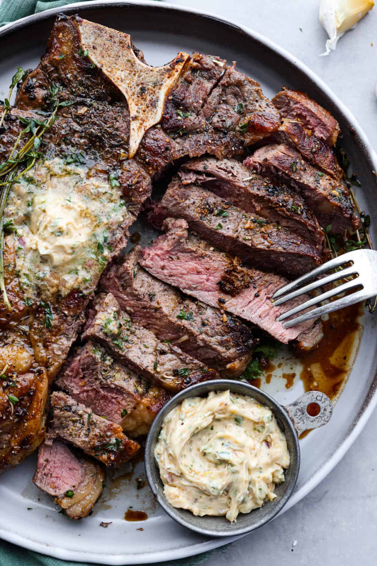 Indulge in the ultimate T-bone steak experience with this recipe! It takes an already awesome cut of meat and cooks it to perfection. The mix of olive oil, spices, and a hot skillet gives you a beautifully seared crust. Top it off with some garlic butter and fresh herbs, then you have the perfect meal ready for any occasion! The meat is cut into slices and served on a gray plate with compound butter.