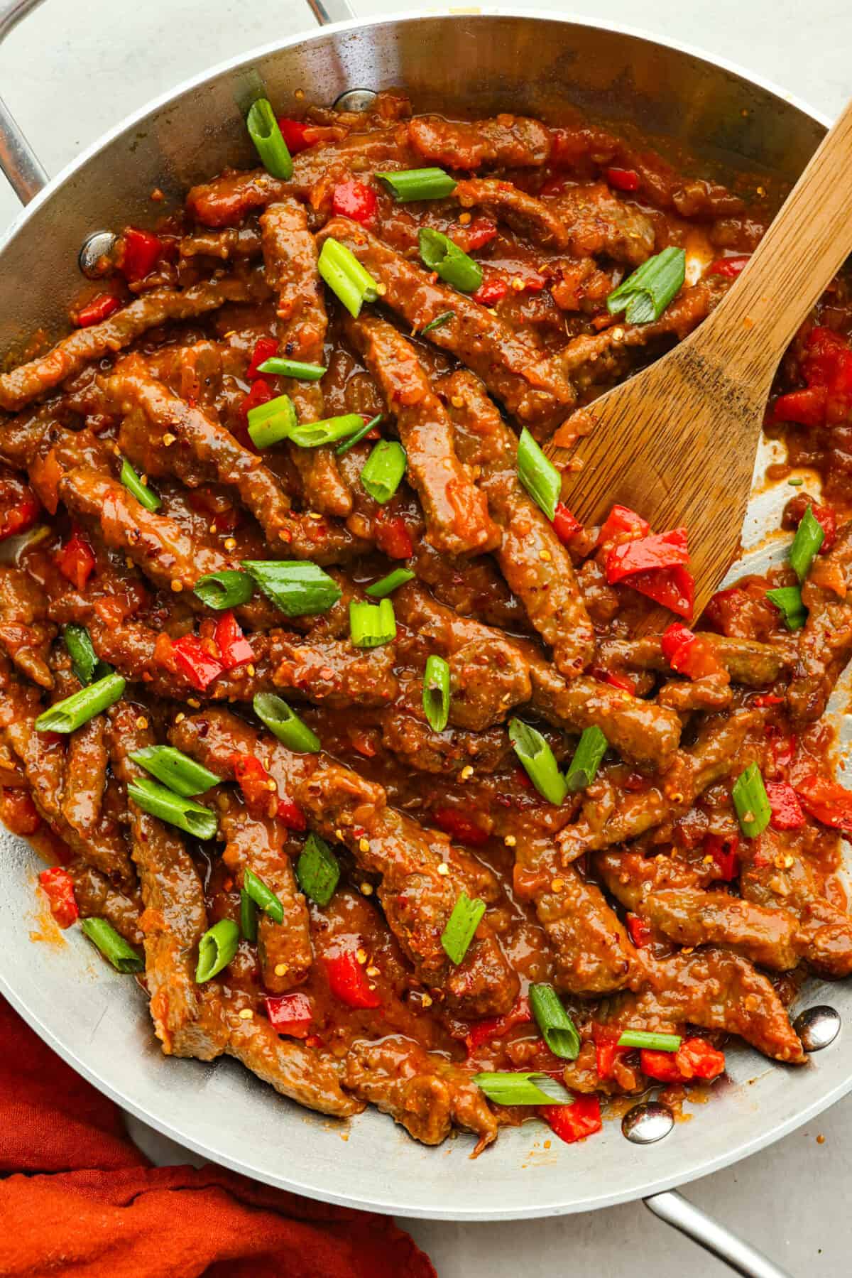Crispy beef strips stir-fried with onion and red bell peppers then tossed in a sweet and tangy sauce. You're going to love this popular Panda Express copycat version of Beijing beef! Top view of Beijing beef in a silver pan with a wooden spoon.