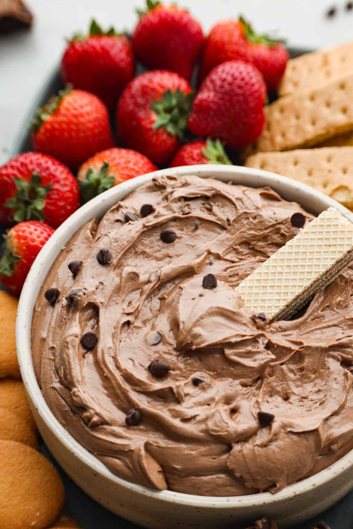 Brownie Batter Dip is an easy, delicious way to get all the brownie flavor without the baking! In this dessert dip recipe, brownie mix, cream cheese, vanilla and whipped topping get combined together for one of the most incredible treats. Serve with cookies, fruit and marshmallows! Hero image of brownie batter dip in a white bowl, served with a wafer cookie.