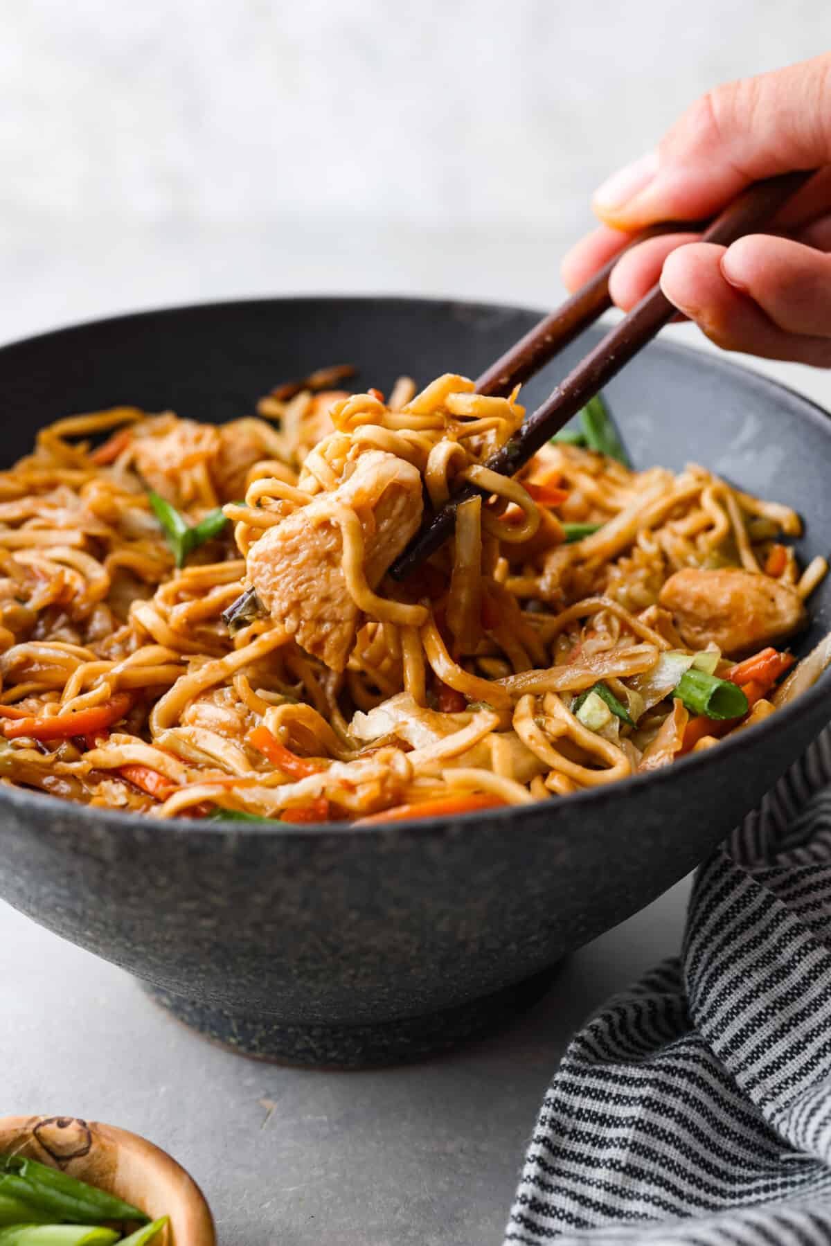 This delicious Chicken Chow Mein is perfect for a quick and easy weeknight meal that your entire family will love! It's made with fresh veggies, noodles, and sautรฉed chicken, tossed in a simple, savory glaze. Picking up a bite of noodles and chicken with chopsticks.