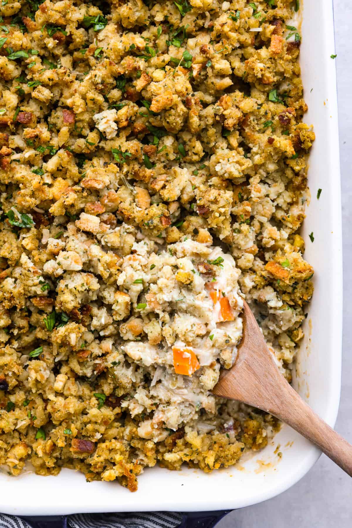 A delicious, hearty casserole made with stuffing mix, shredded chicken, vegetables, and all the tasty seasonings. Chicken and stuffing casserole is a fun twist on a classic dish and the perfect addition to your Thanksgiving lineup! Closeup of a scoop of chicken and stuffing casserole on a wooden spoon.
