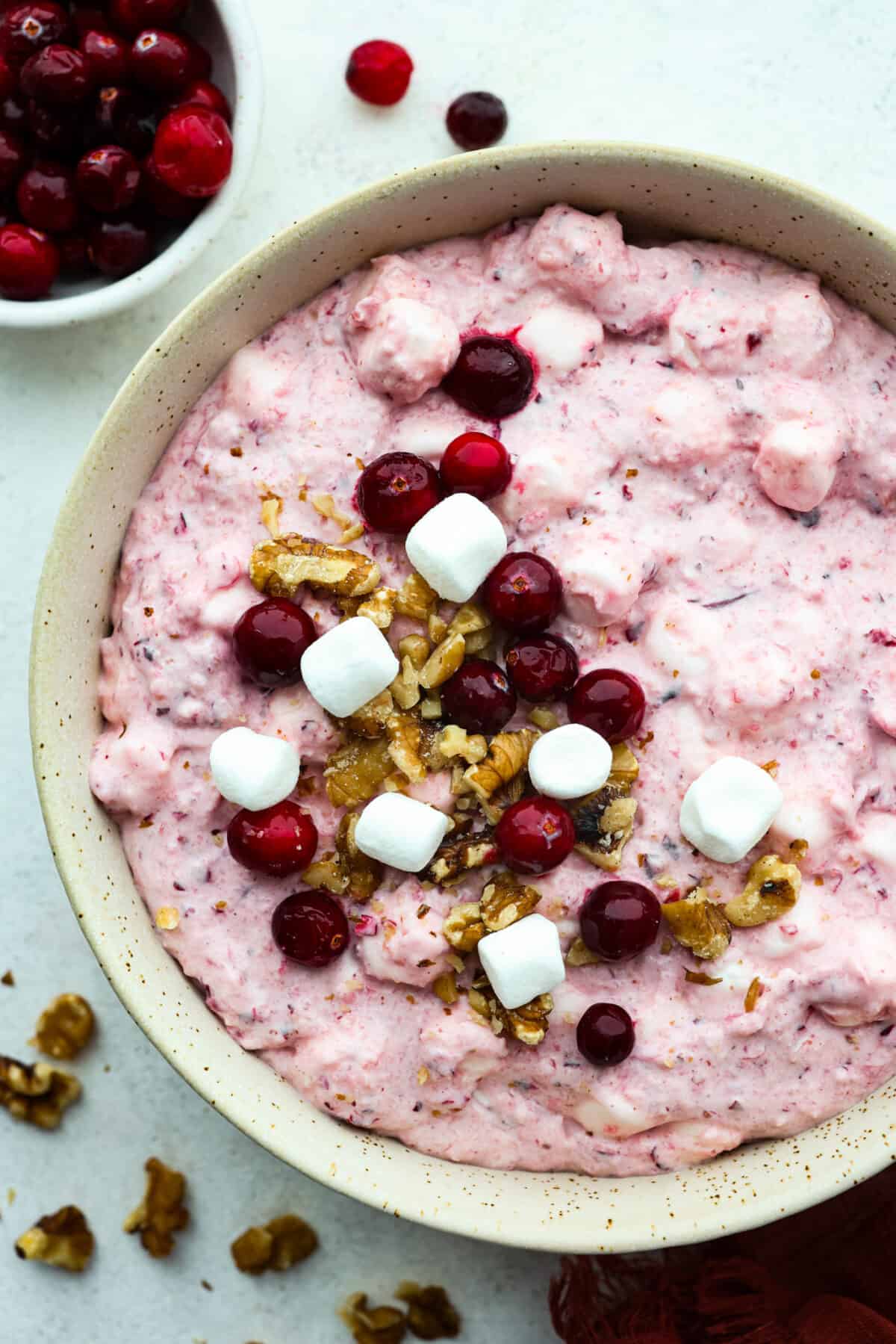 Cranberry Cheesecake Fluff is aย creamy and delicious fluff salad made with cranberries and walnuts. It is so easy to make and a MUST for the holidays! Top-down view of cranberry fluff salad served in a white bowl, topped with marshmallows, cranberries, and chopped walnuts.