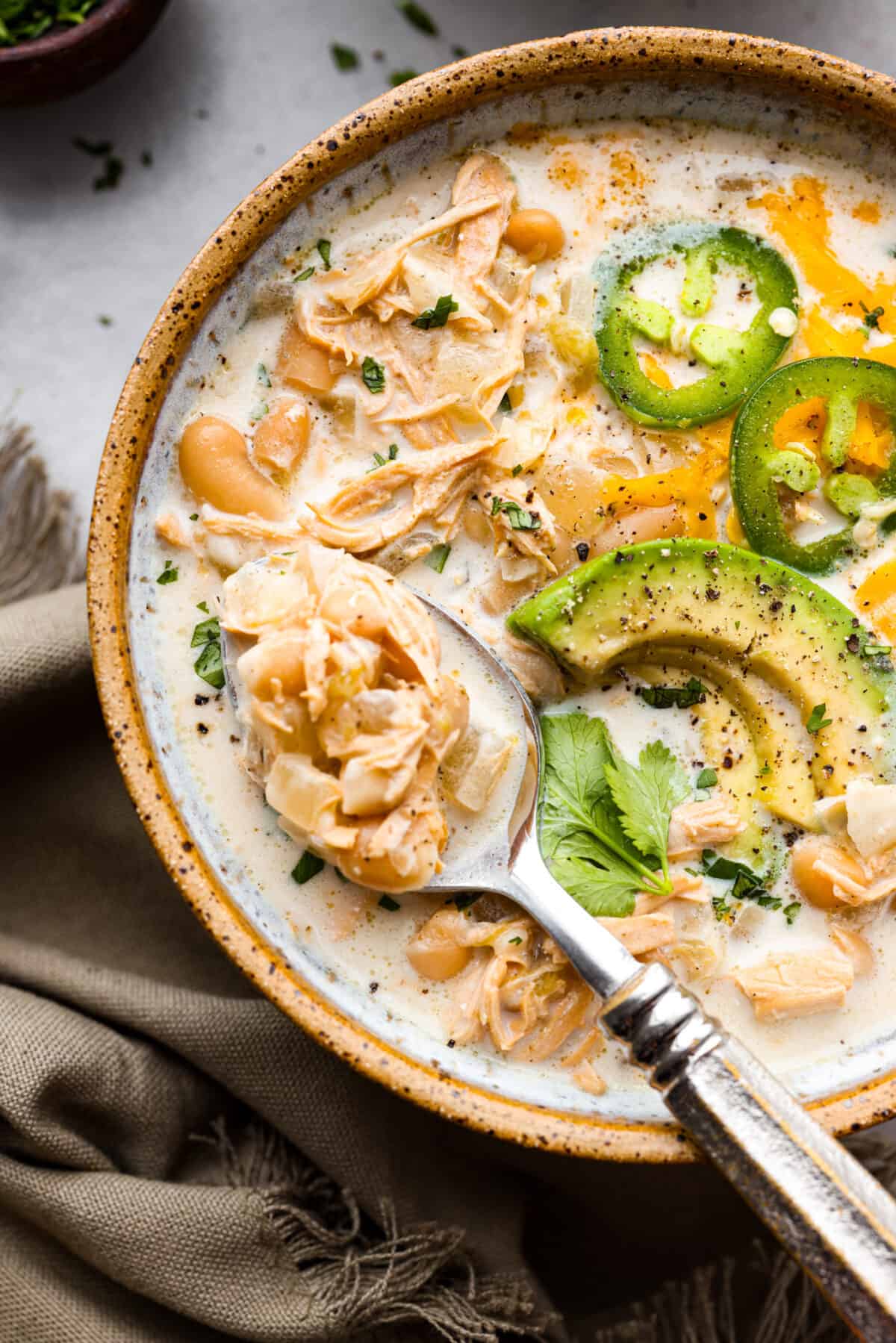 Crockpot White Chicken Chili combines chopped onions, garlic, spices, rotisserie chicken, green chiles, hearty beans, and chicken broth. The best part is the flavors meld in your slow cooker while it cooks all day. A close up of a bowl of white chicken chili with a spoon and garnished with some jalapeños, avocado slices and some cilantro.