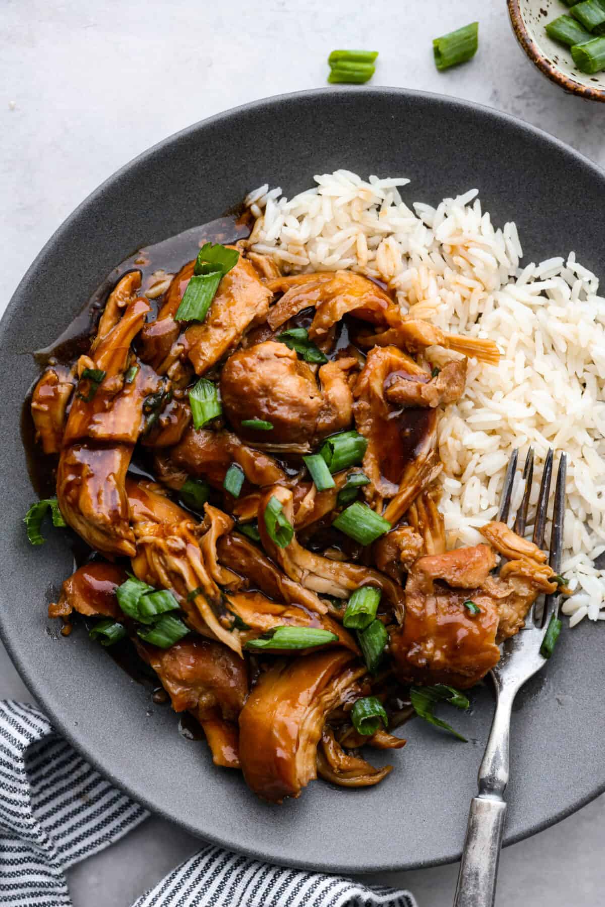 Tender and juicy chicken thighs simmered in a brown sugar bourbon sauce all day long. Your family will love this melt-in-your-mouth crockpot bourbon chicken. A classic New Orleans recipe made easy in the crockpot! Close top view of shredded crockpot bourbon chicken on a gray plate with rice.