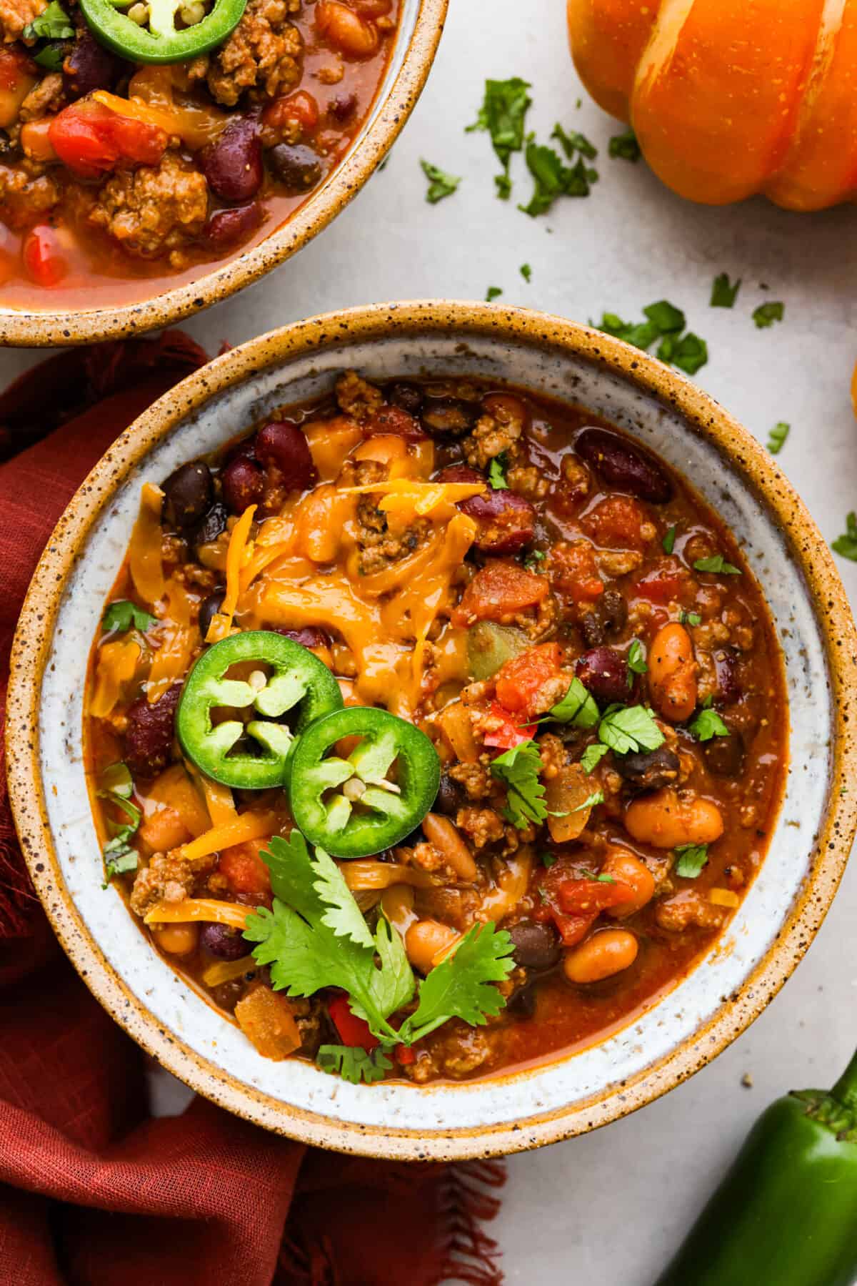 Pumpkin chili is the perfect hearty meal for cold weather. It features perfectly seasoned ground beef, tender beans, and velvety pumpkin puree. Each bowl is packed with fall flavor, making it a cozy and delicious choice for chilly days. Overhead shot of a bowl of pumpkin chili topped with jalapeรฑos, cheese and cilantro.
