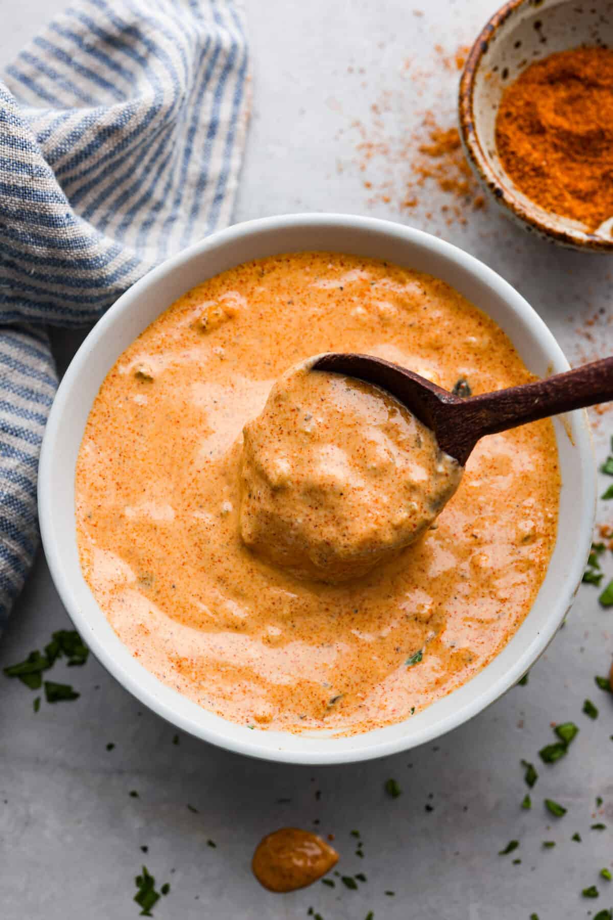Remoulade sauce, a tasty dip from Louisiana, is made with mayonnaise, mustard, hot sauce, relish, and some Cajun spices for the best flavor ever! Whether you use it as a dip or spread, you're going to love the kick of flavor it adds to your favorite recipes. Remoulade sauce in a white bowl, being mixed with a wooden bowl.
