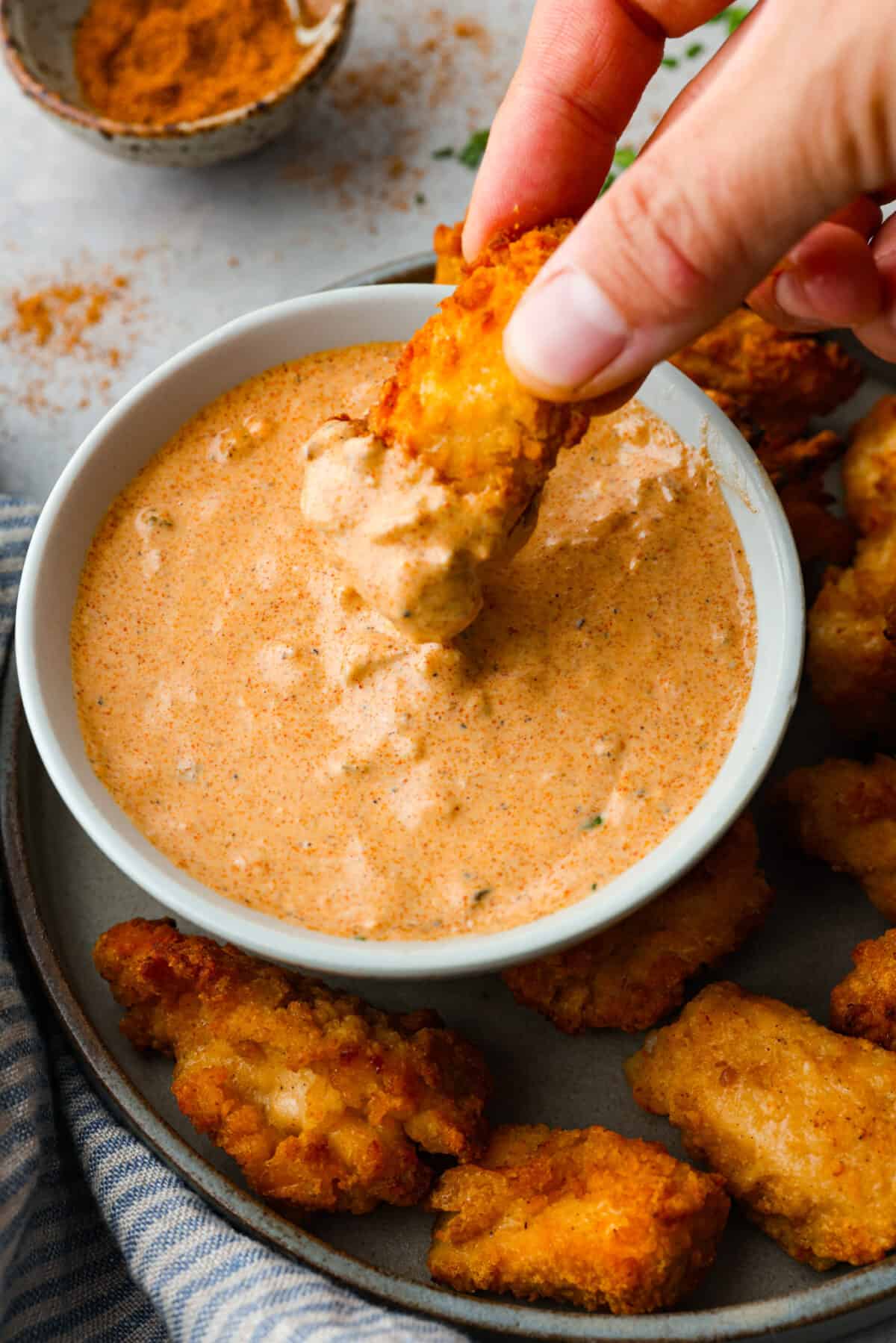 Remoulade sauce, a tasty dip from Louisiana, is made with mayonnaise, mustard, hot sauce, relish, and some Cajun spices for the best flavor ever! Whether you use it as a dip or spread, you're going to love the kick of flavor it adds to your favorite recipes. Dipping chicken into the sauce.