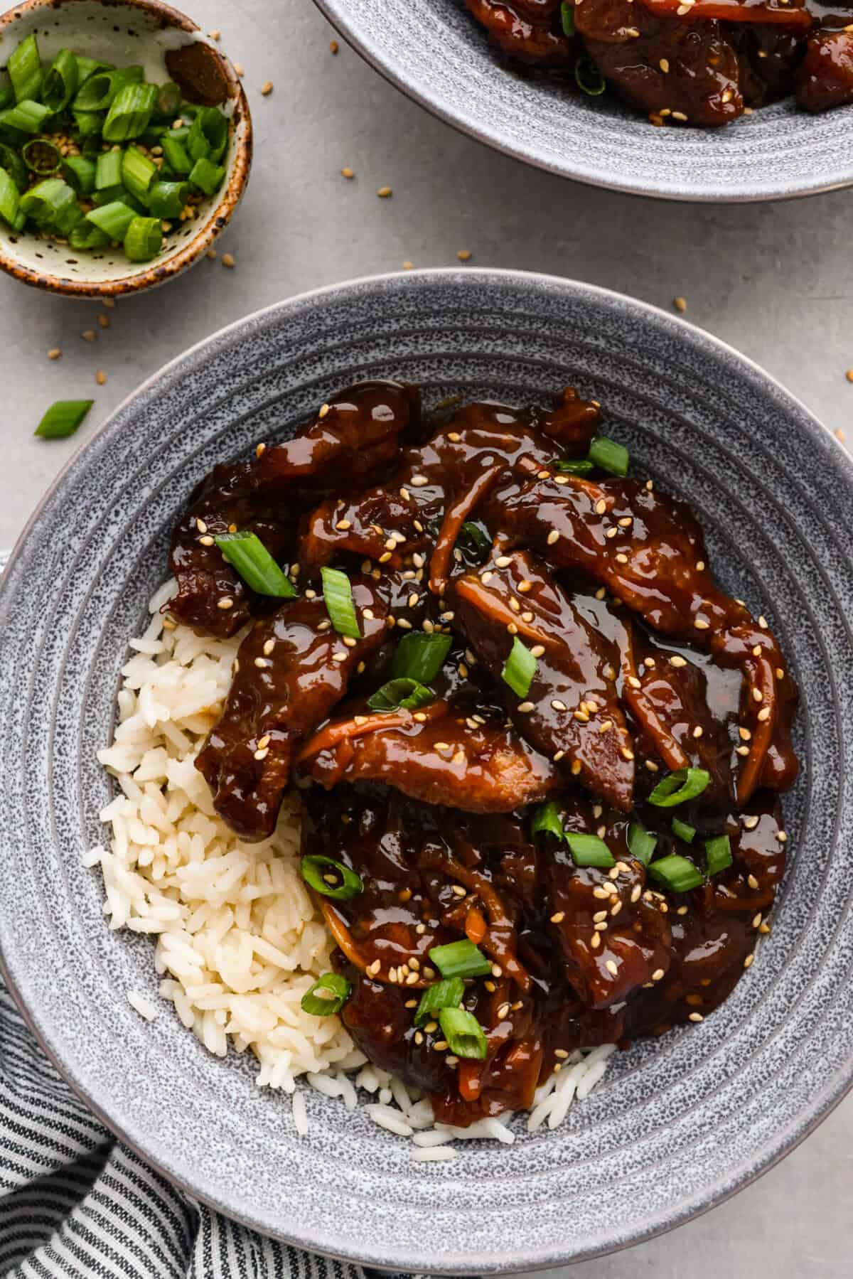 Tender pieces of flank steak are slow-cooked in the tastiest sweet and savory sauce. This slow cooker Mongolian beef is melt-in-your-mouth perfection and bursting with flavor. Quick and easy to prepare and one of our favorite slow cooker meals! Overhead view of slow cooker Mongolian beef in a gray bowl over rice. Green onions and sesame seeds garnished on top.