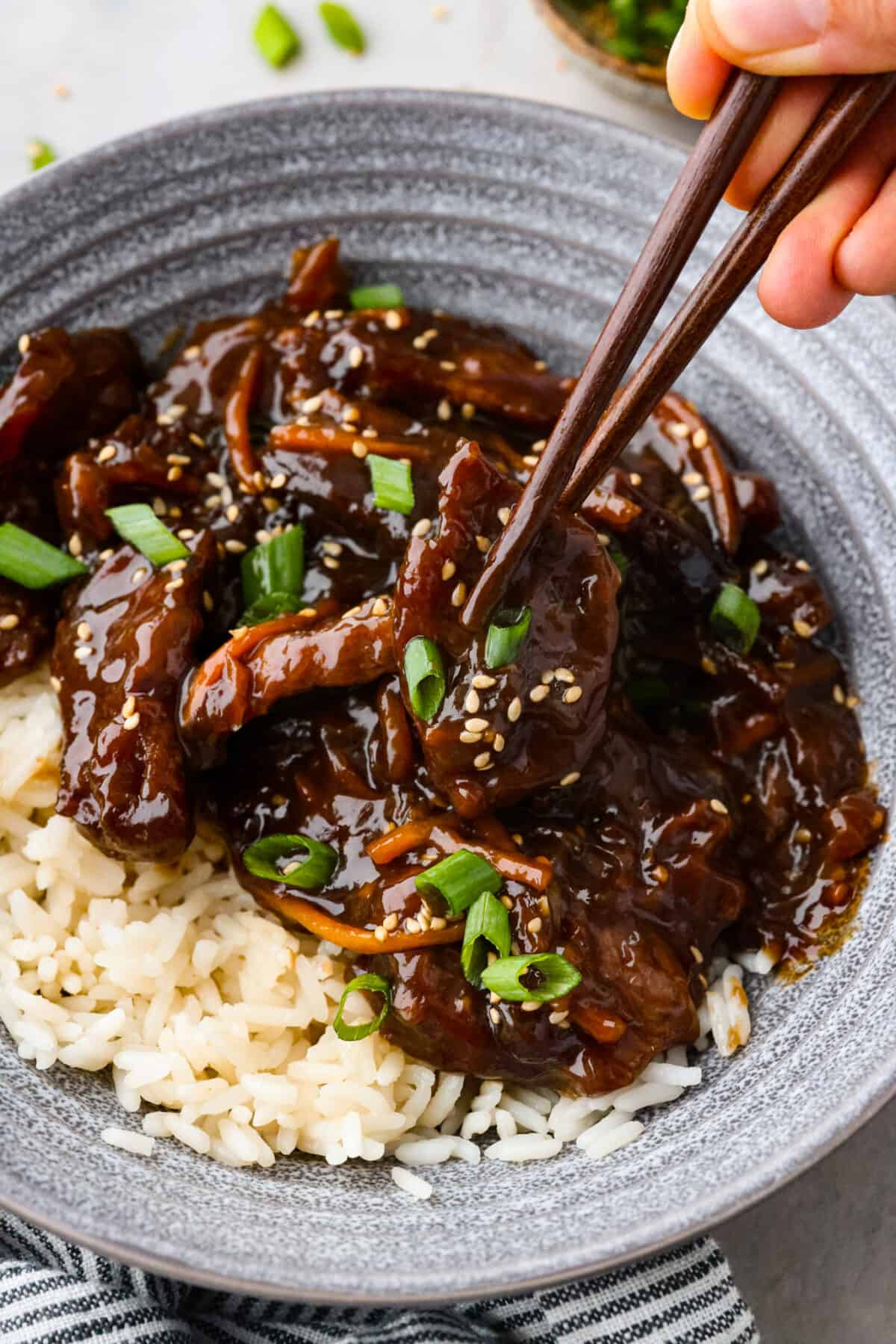 Tender pieces of flank steak are slow-cooked in the tastiest sweet and savory sauce. This slow cooker Mongolian beef is melt-in-your-mouth perfection and bursting with flavor. Quick and easy to prepare and one of our favorite slow cooker meals! Close view of chopsticks lifting up beef from a bowl of slow cooker Mongolian beef over rice.