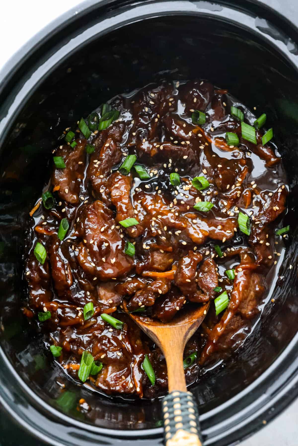Tender pieces of flank steak are slow-cooked in the tastiest sweet and savory sauce. This slow cooker Mongolian beef is melt-in-your-mouth perfection and bursting with flavor. Quick and easy to prepare and one of our favorite slow cooker meals! Top view of slow cooker mongolian beef in a black crock pot. A wooden spoon is in the crockpot.