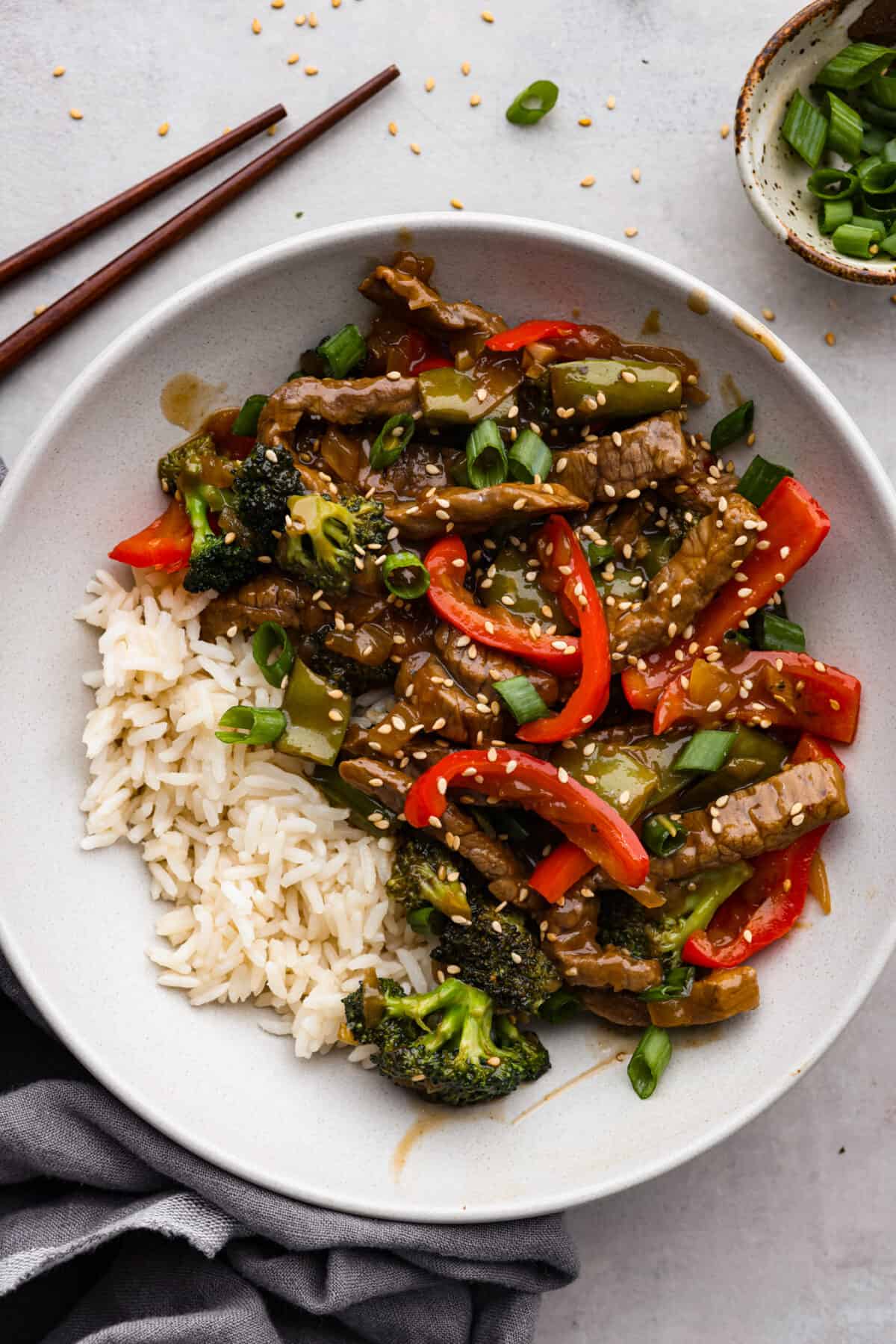 Delicious Szechuan Beef is filled with mouthwatering bold flavor! Tender strips of beef, bell peppers, snap peas, and broccoli are stir-fried together with a perfectly sweet, spicy, tangy sauce. It's way better than takeout and cooks up in under 20 minutes! Szechuan beef and veggies served with rice in a white bowl.