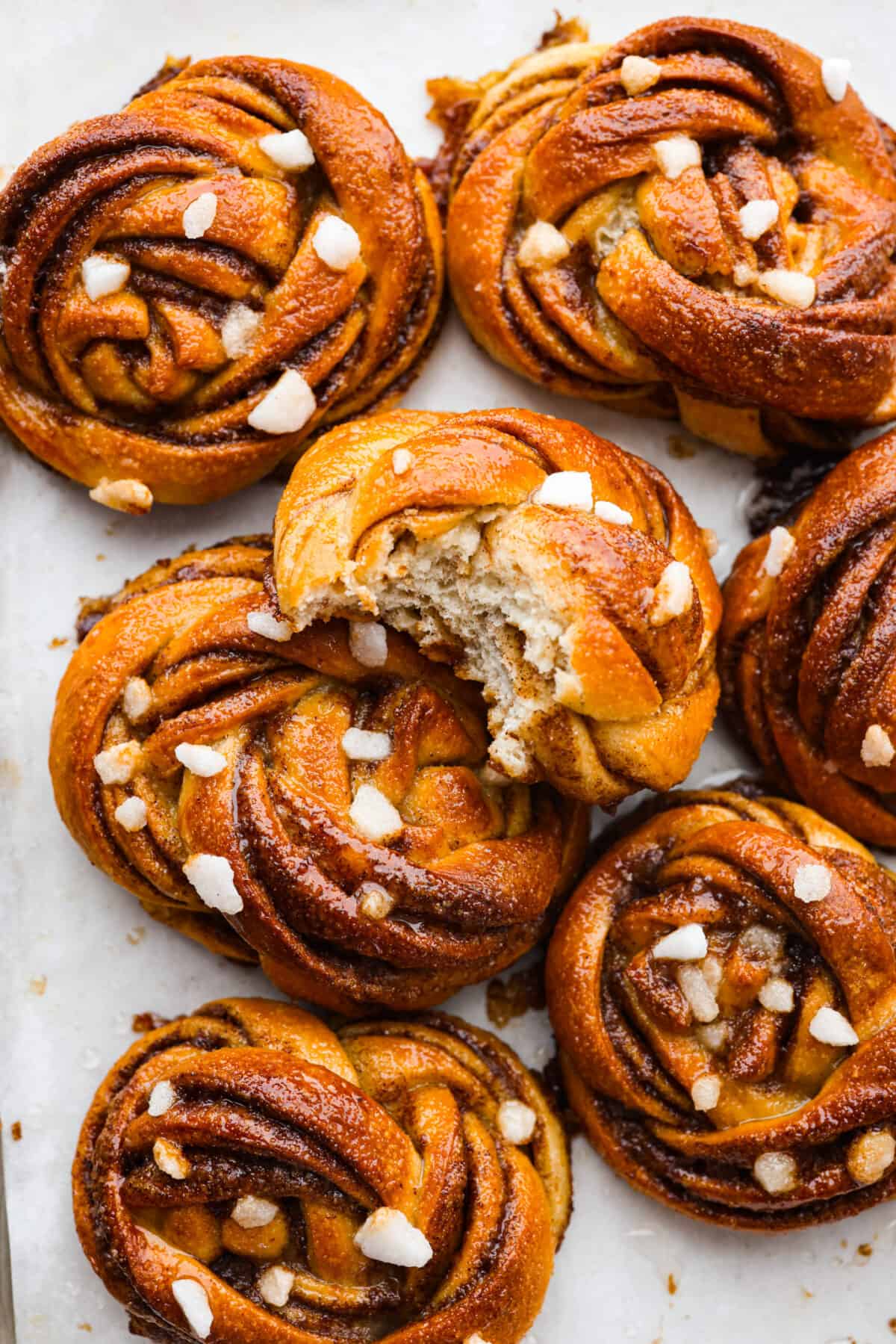These Swedish cinnamon buns, also known as Kanelbullar, are just the dessert to make if you’re craving something soft, sweet, and spiced to perfection! You’re not going to be able to resist going in for another bite, especially if they’re fresh out of the oven! Closeup of kanelbullar buns topped with pearl sugar. One has been torn in half.