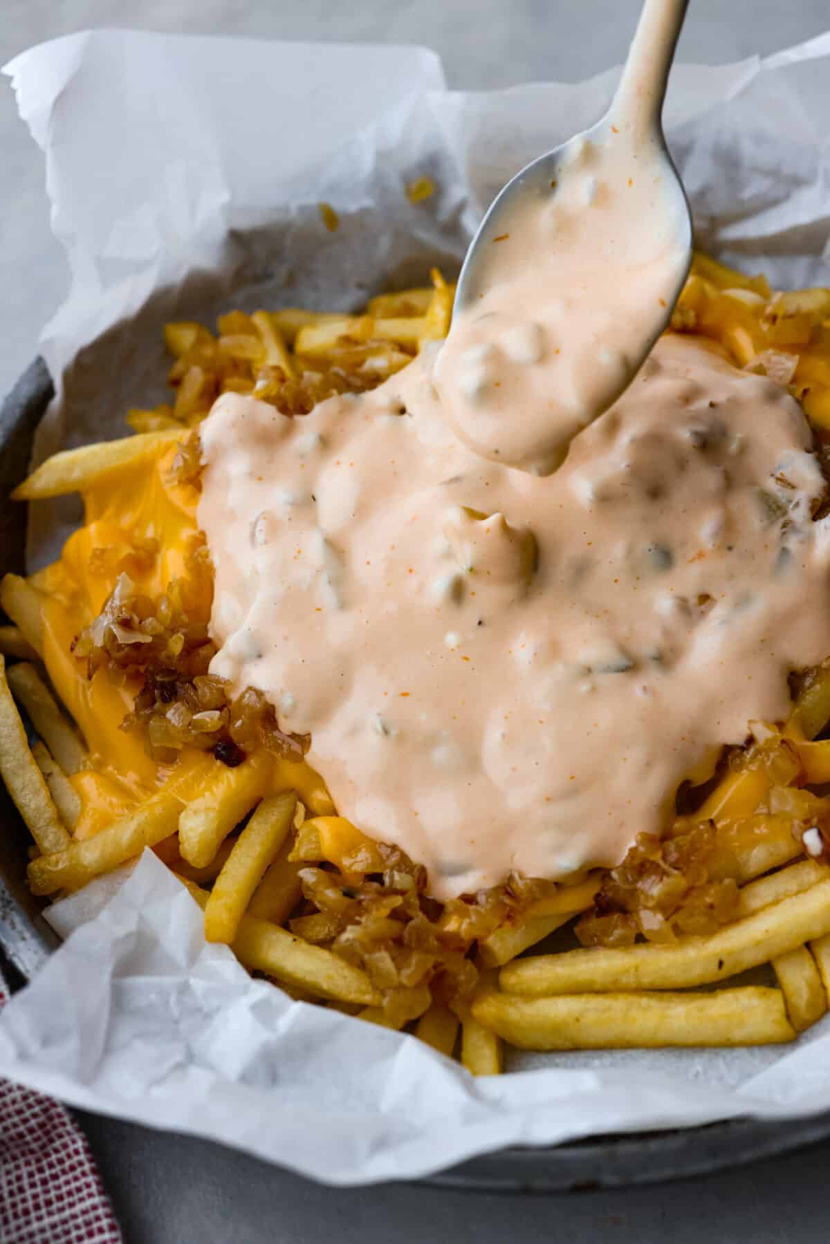 Skip the takeout and make your Animal Fries at home! Inspired by In-N-Out Burger, this recipe features crispy, golden fries piled high with melted American cheese, caramelized onions, and homemade animal sauce! (A tasty blend of ketchup, relish, vinegar, and mayo!) Spooning the zesty sauce over the animal fries.