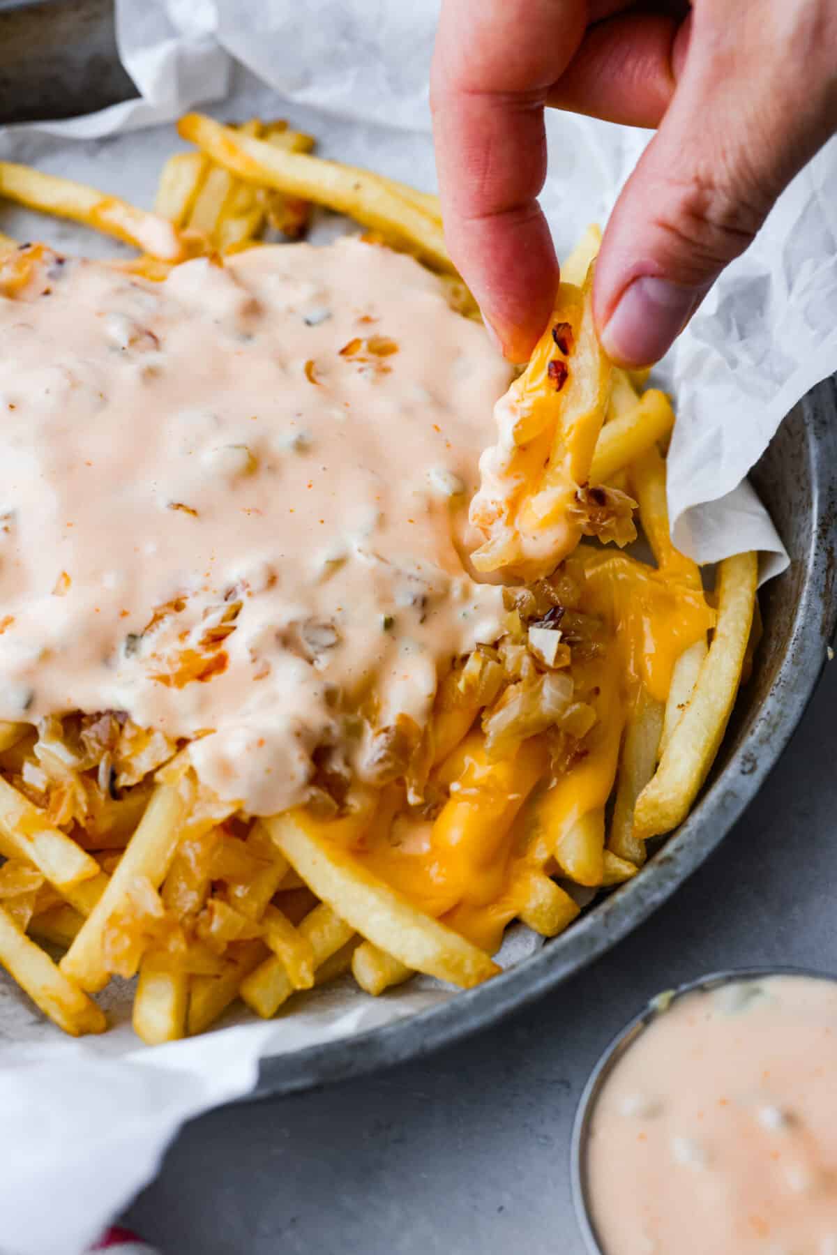 Skip the takeout and make your Animal Fries at home! Inspired by In-N-Out Burger, this recipe features crispy, golden fries piled high with melted American cheese, caramelized onions, and homemade animal sauce! (A tasty blend of ketchup, relish, vinegar, and mayo!) 2 fries being picked up.