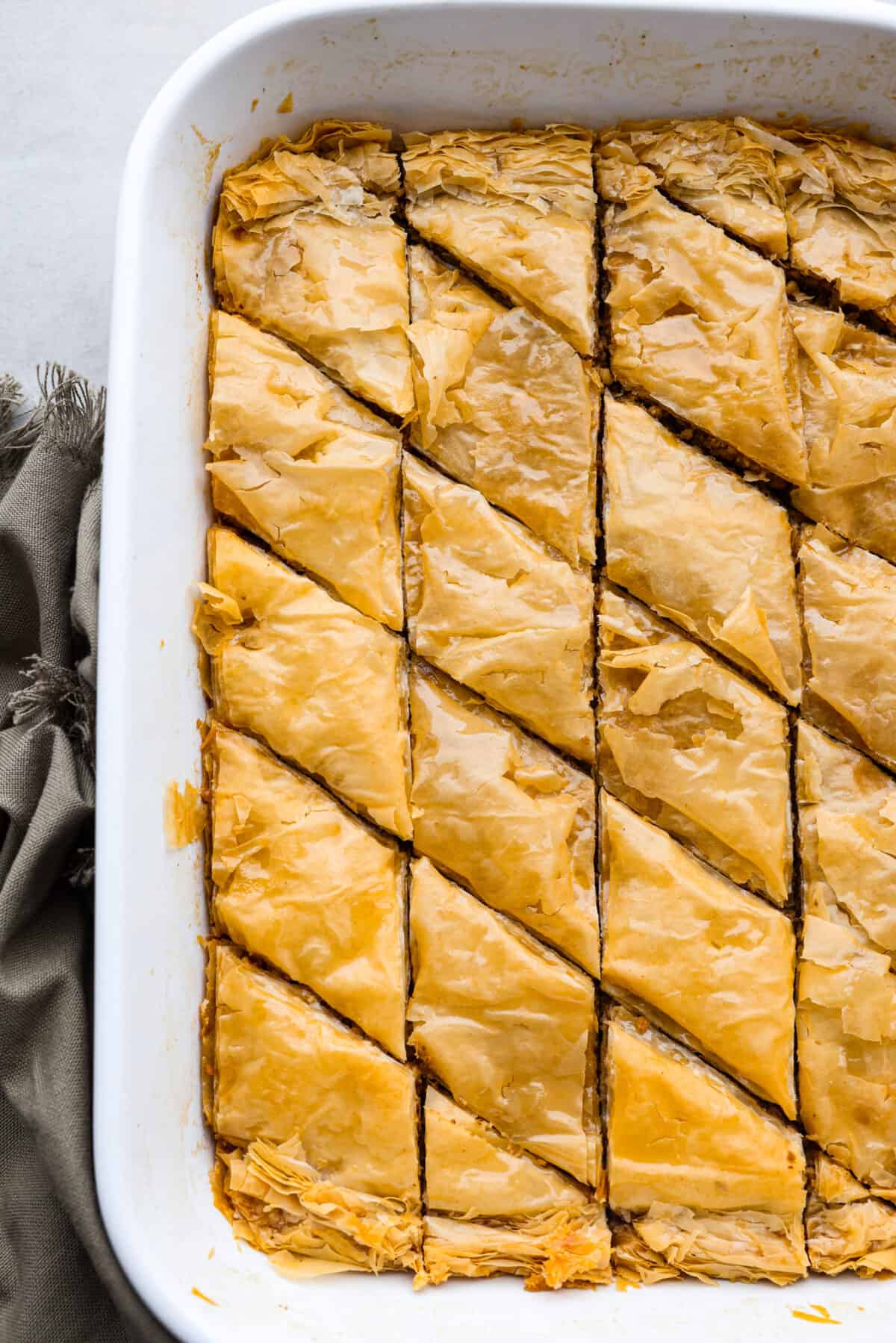 Phyllo pastry and walnuts are layered together then baked until the pastry is deliciously flaky. A sweet honey syrup is poured over the top which soaks into the baklava and makes every bite heavenly. Top-down view of baklava in a white baking dish.
