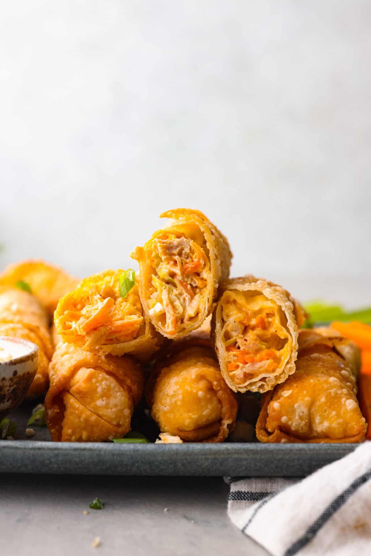 Wait until you taste these Buffalo Chicken Egg Rolls' crispy texture and bold flavors! A delicious mixture of cream cheese, hot sauce, and spices envelops shredded chicken, carrots, and cabbage, creating a perfect balance of crunch and zing. Egg rolls stacked on one another. The top two have been cut in half so that you can see the inside.