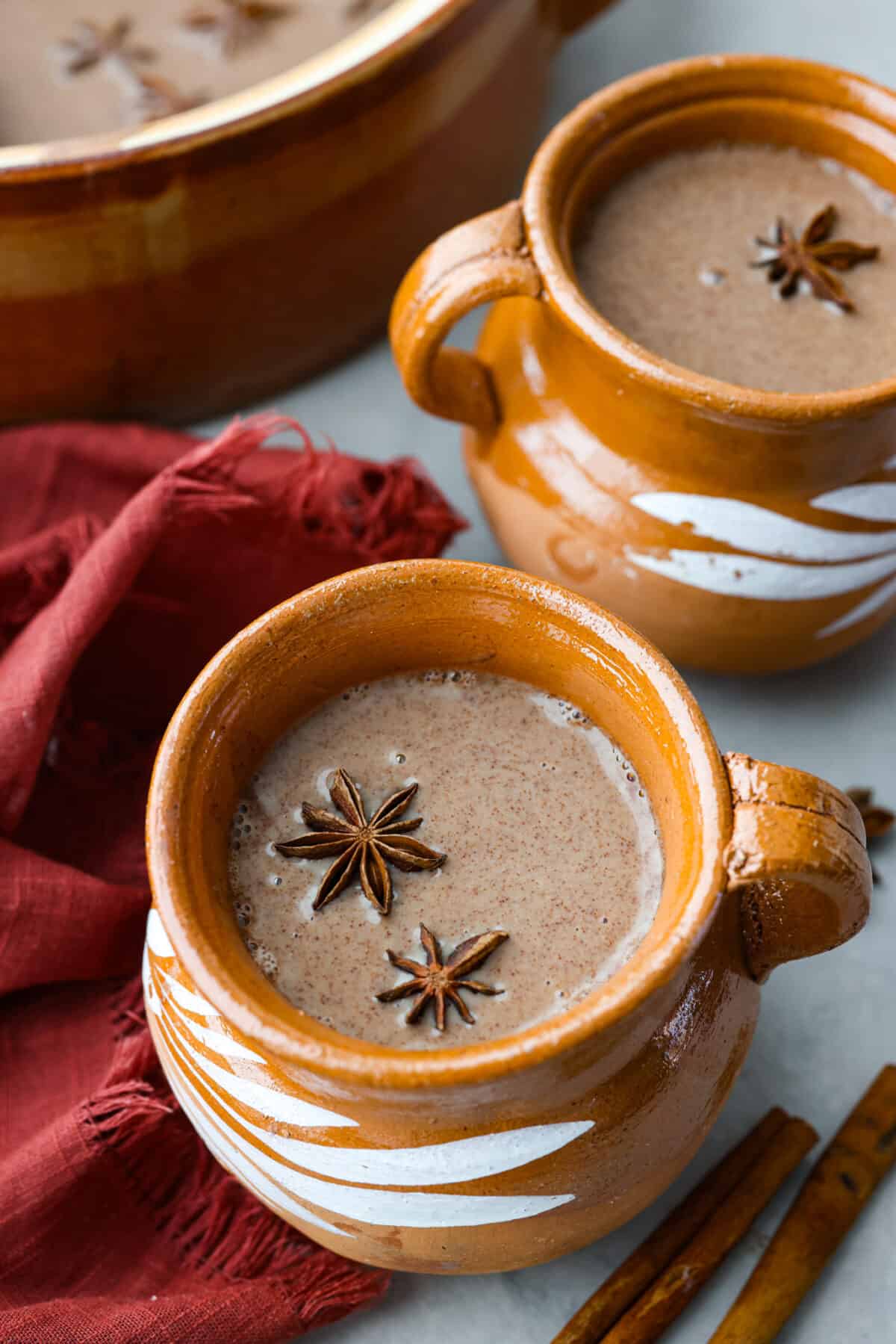 Get toasty from the inside out with champurrado, a delicious variation of Mexican hot chocolate! Deep, melty chocolate meets warming spices and brown sugar, all thickened with corn flour for a velvety smooth sip. Perfect for chasing any winter blues away! Champurrado in a brown mug. It's filled with star anise and surrounded with cinnamon sticks.