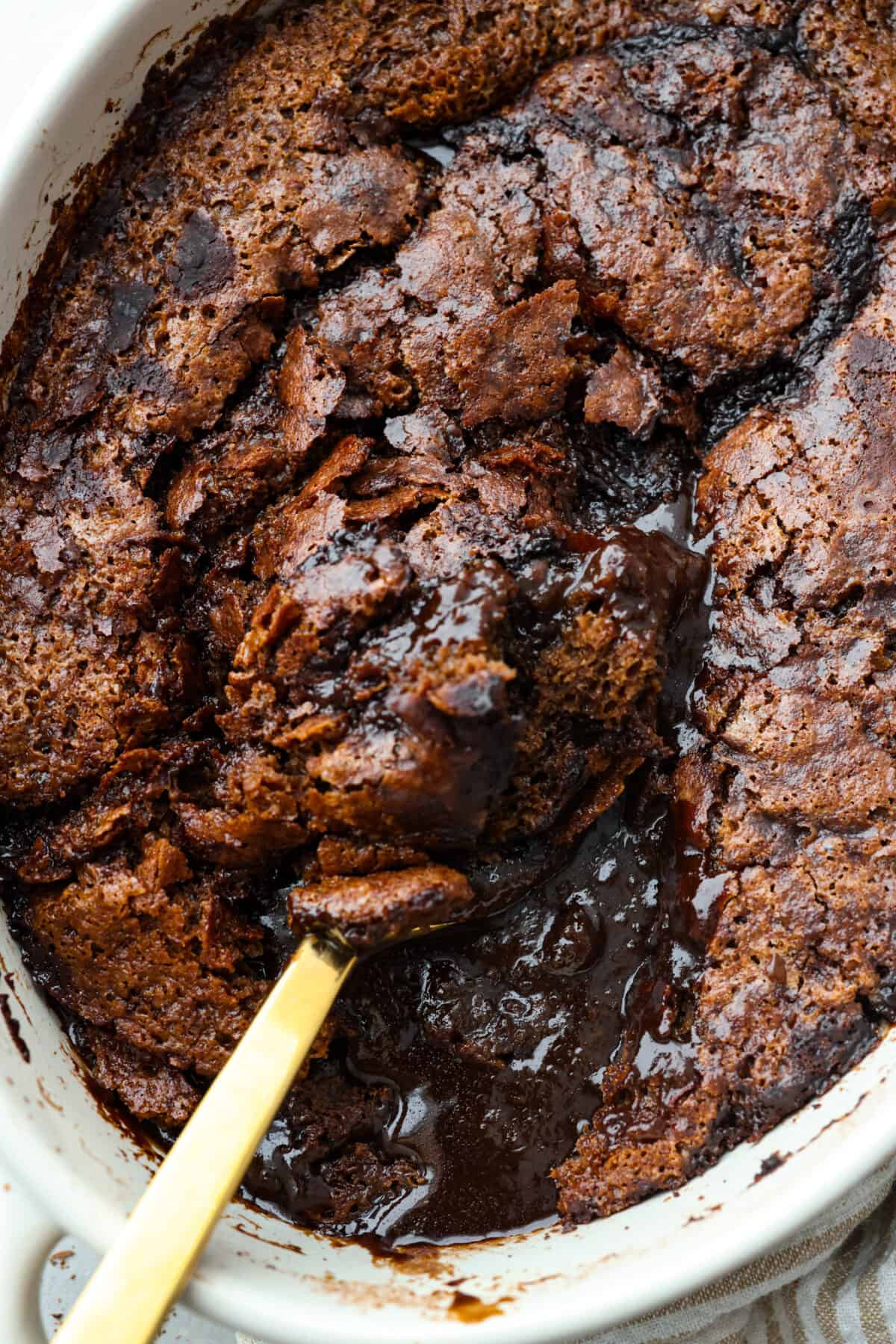 Everyone will want to eat dessert first with this Chocolate Cobbler โ a simple dessert that's all about gooey chocolate goodness. The velvety inside and crispy top create a perfect combo for a satisfying, easy-to-make treat! a close up of chocolate cobbler in a pan with a large, gold serving spoon.