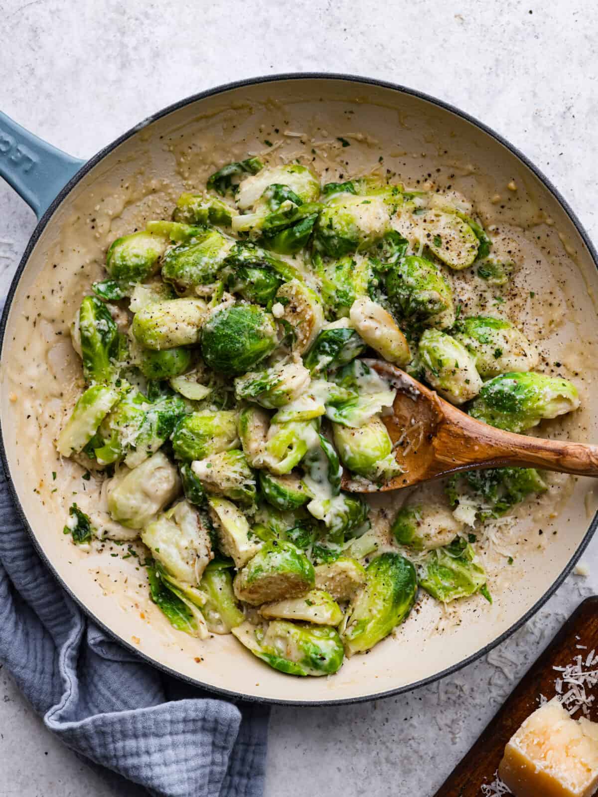 Creamy parmesan garlic Brussels sprouts sauteed and smothered in a rich and cheesy parmesan garlic sauce. Elevate your Brussels sprouts with this easy and delicious side dish! Top view of creamy brussels sprouts in a skillet with a wooden spoon.