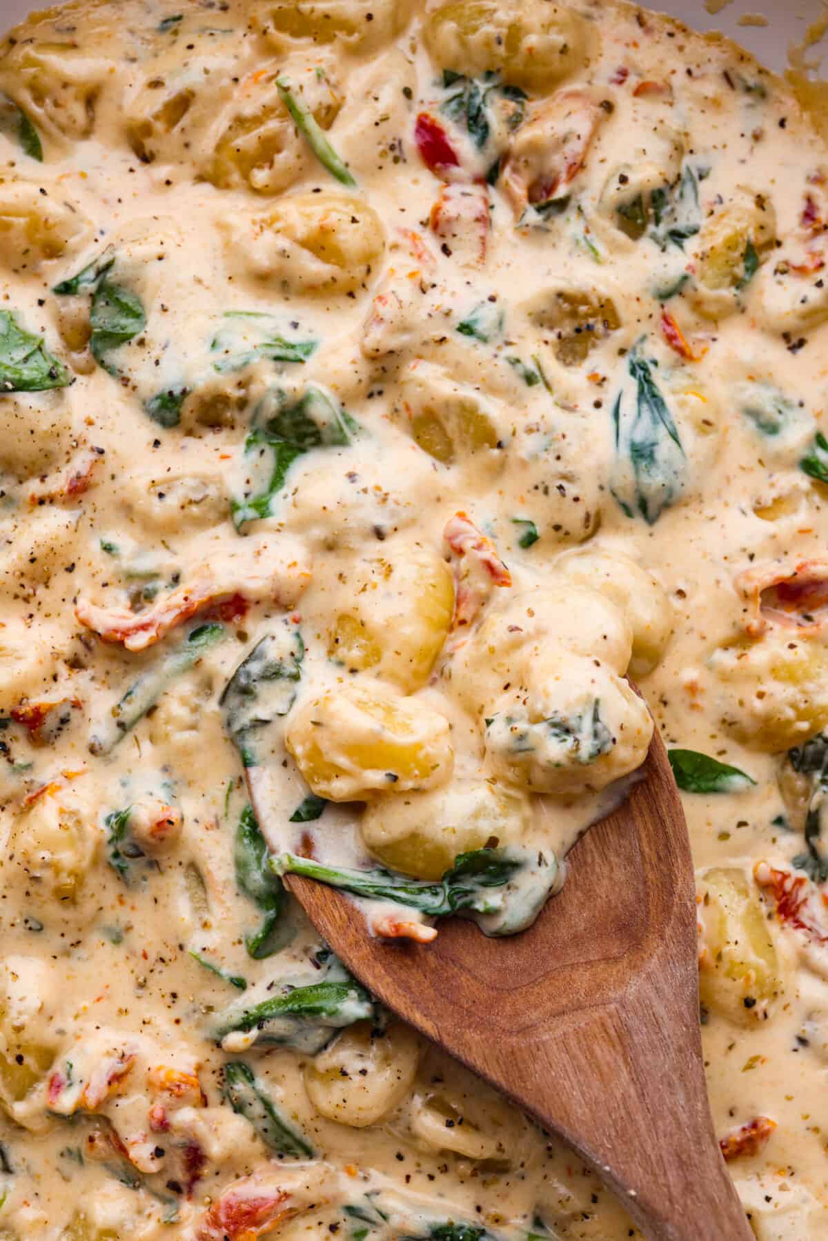 Creamy Tuscan Parmesan Gnocchi has the most incredible creamy sauce with spinach and sun-dried tomatoes. This is an easy comforting meal packed with flavor that can be on the dinner table in less than 30 minutes! Closeup of the gnocchi in a creamy sun-dried tomato sauce. It is being stirred with a wooden spoon.