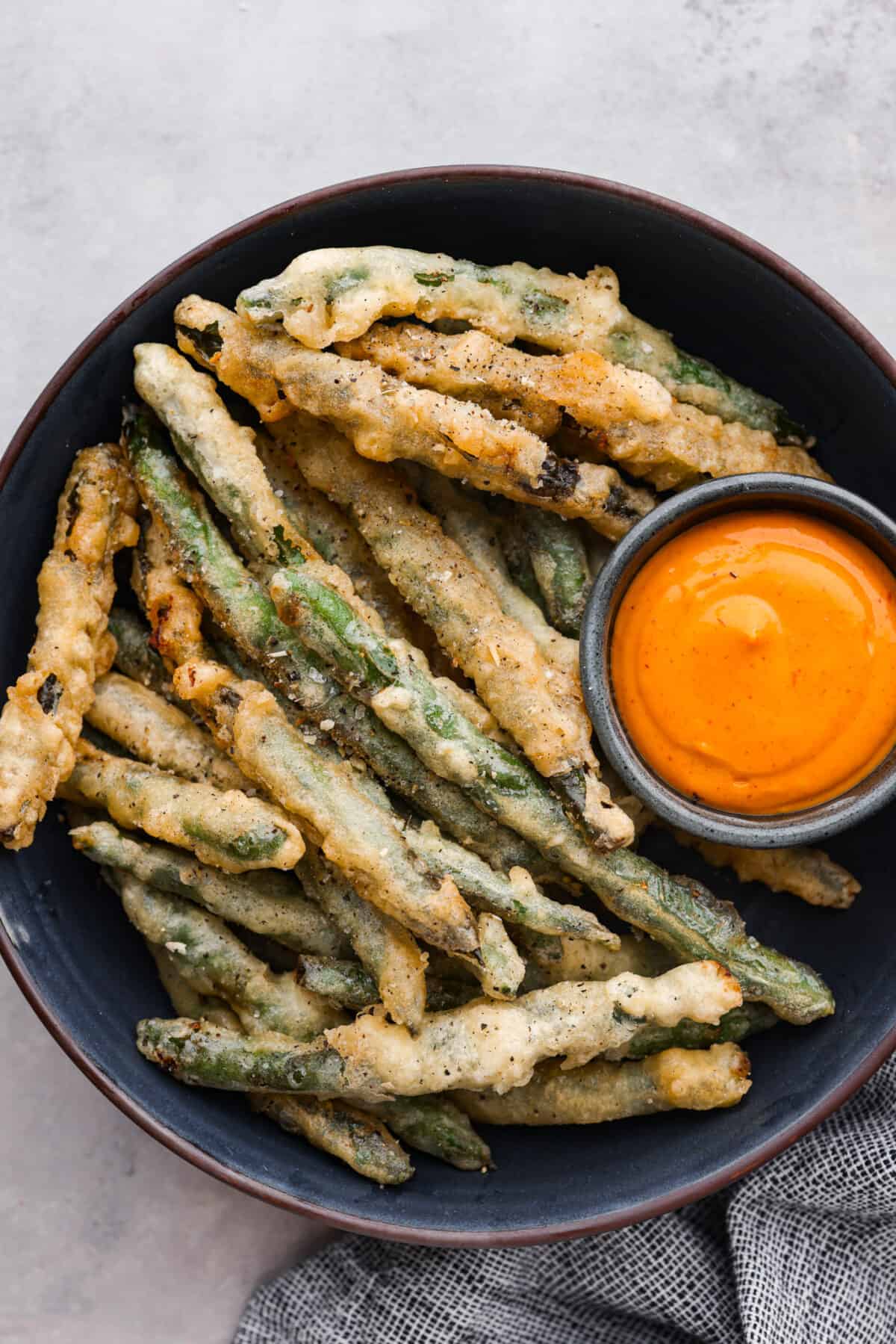 One of the best things on the menu at P.F. Changs is their Crispy Fried Green Beans. These green beans taste even better, and you can make them at home! They have a delightful crunch and are seasoned to perfection! A bowl of fried green beans with a small bowl of orange dipping sauce.