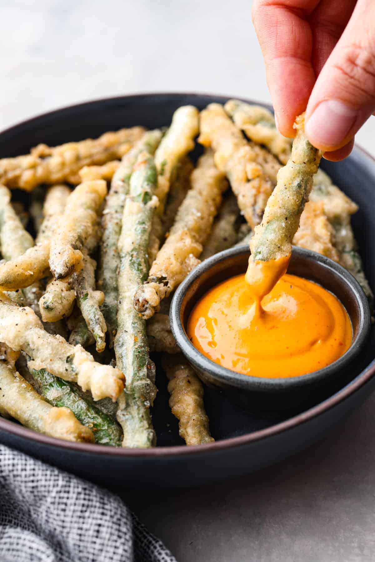 One of the best things on the menu at P.F. Changs is their Crispy Fried Green Beans. These green beans taste even better, and you can make them at home! They have a delightful crunch and are seasoned to perfection! A crispy green bean being dipped into some orange sauce.