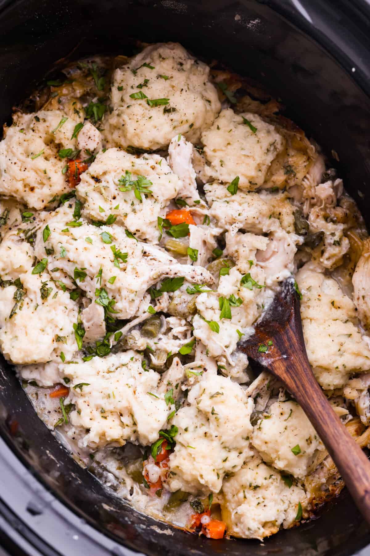 Crockpot chicken and dumplings...the ultimate comfort meal ready when you get home! Tender chicken and vegetables topped with homemade dumplings. Easy to assemble and slow-cooked to perfection. Top view of crockpot chicken and dumplings in a crockpot with a wooden spoon.