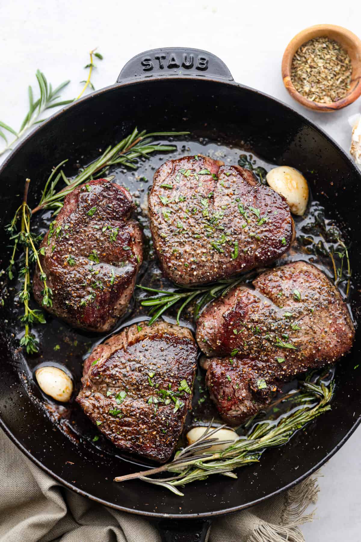 Juicy and succulent filet mignon is perfect for dinner or any special occasion! Tender steak is seasoned, seared, and basted with garlic herb butter. This is a steakhouse-quality recipe that is so easy to make at home! Top view of filet mignon in a black cast iron pan with garlic and herbs.
