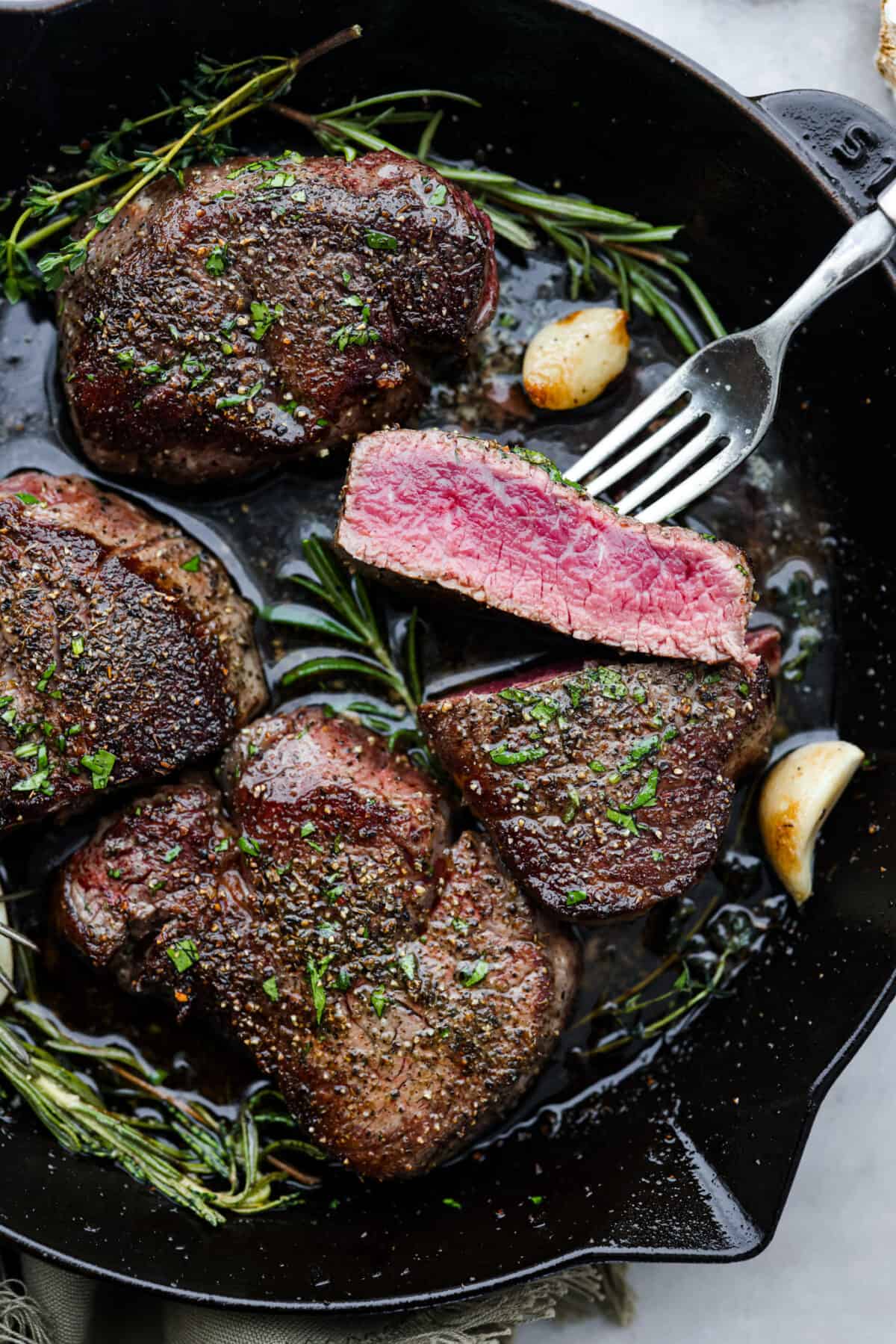Juicy and succulent filet mignon is perfect for dinner or any special occasion! Tender steak is seasoned, seared, and basted with garlic herb butter. This is a steakhouse-quality recipe that is so easy to make at home! Top view of filet mignon in a black cast iron skillet with herbs and garlic.
