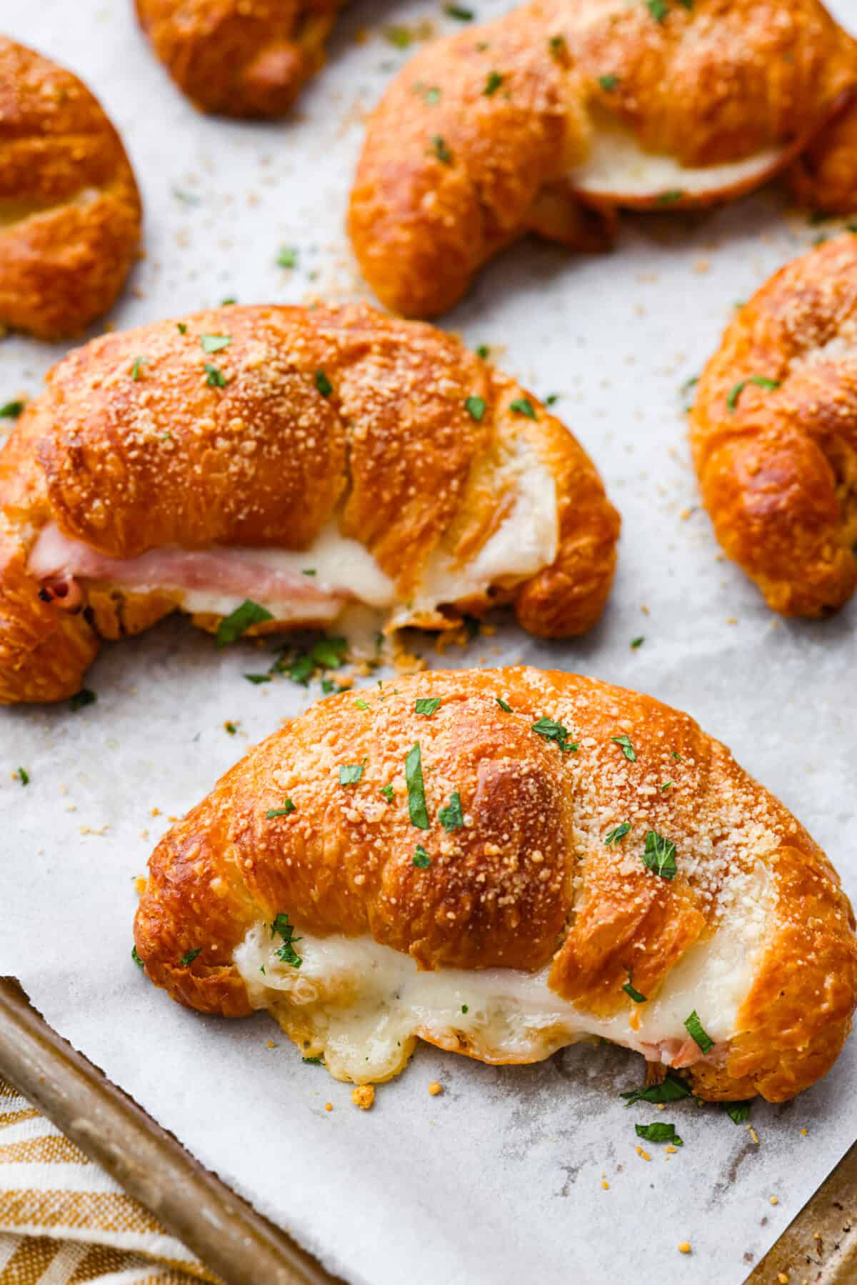 Get ready to devour the BEST ham and cheese croissant! Buttery warm croissants filled with a combination of melty cheeses and thinly sliced ham. These easy-to-make croissant sandwiches are perfect any time of day. Close view of ham and cheese croissants on a baking sheet pan garnished with fresh parsley.