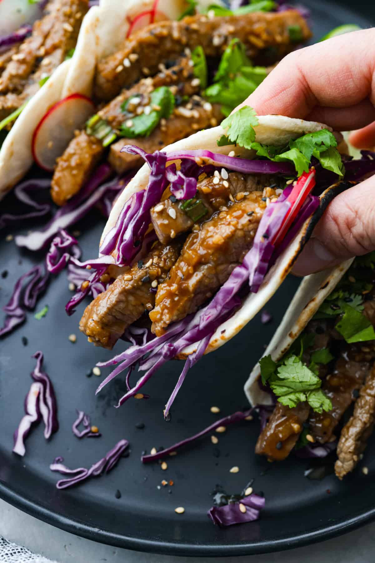 Enjoy the fusion of Asian flavors in these tasty Korean tacos! Marinated beef bulgogi nestled in soft tortillas and topped with cabbage, cilantro, radishes, and sesame seeds. Close view of a hand holding a taco.