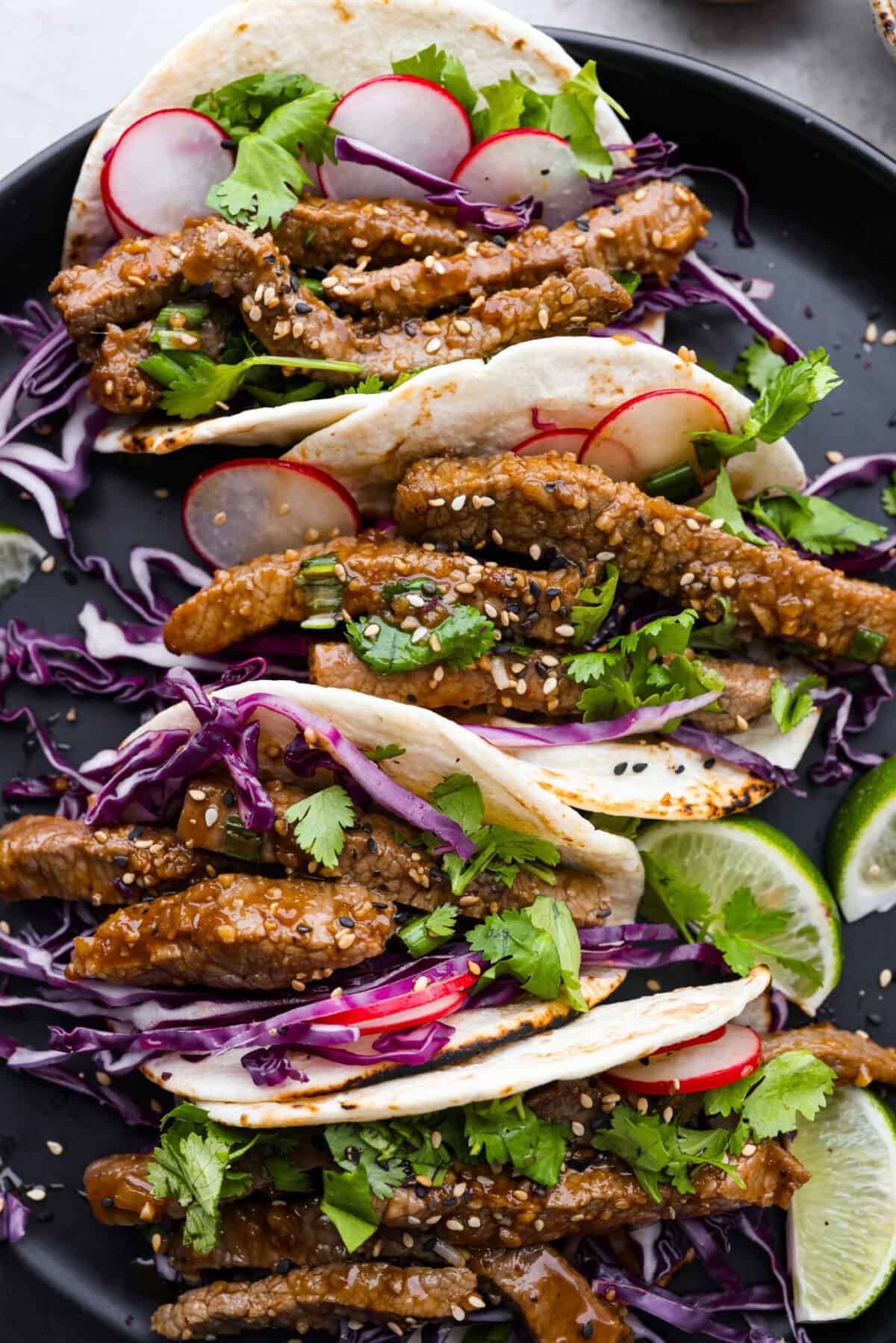 Enjoy the fusion of Asian flavors in these tasty Korean tacos! Marinated beef bulgogi nestled in soft tortillas and topped with cabbage, cilantro, radishes, and sesame seeds. Close view of tacos on a black plate garnished with cabbage, cilantro, radishes, limes, and sesame seeds.