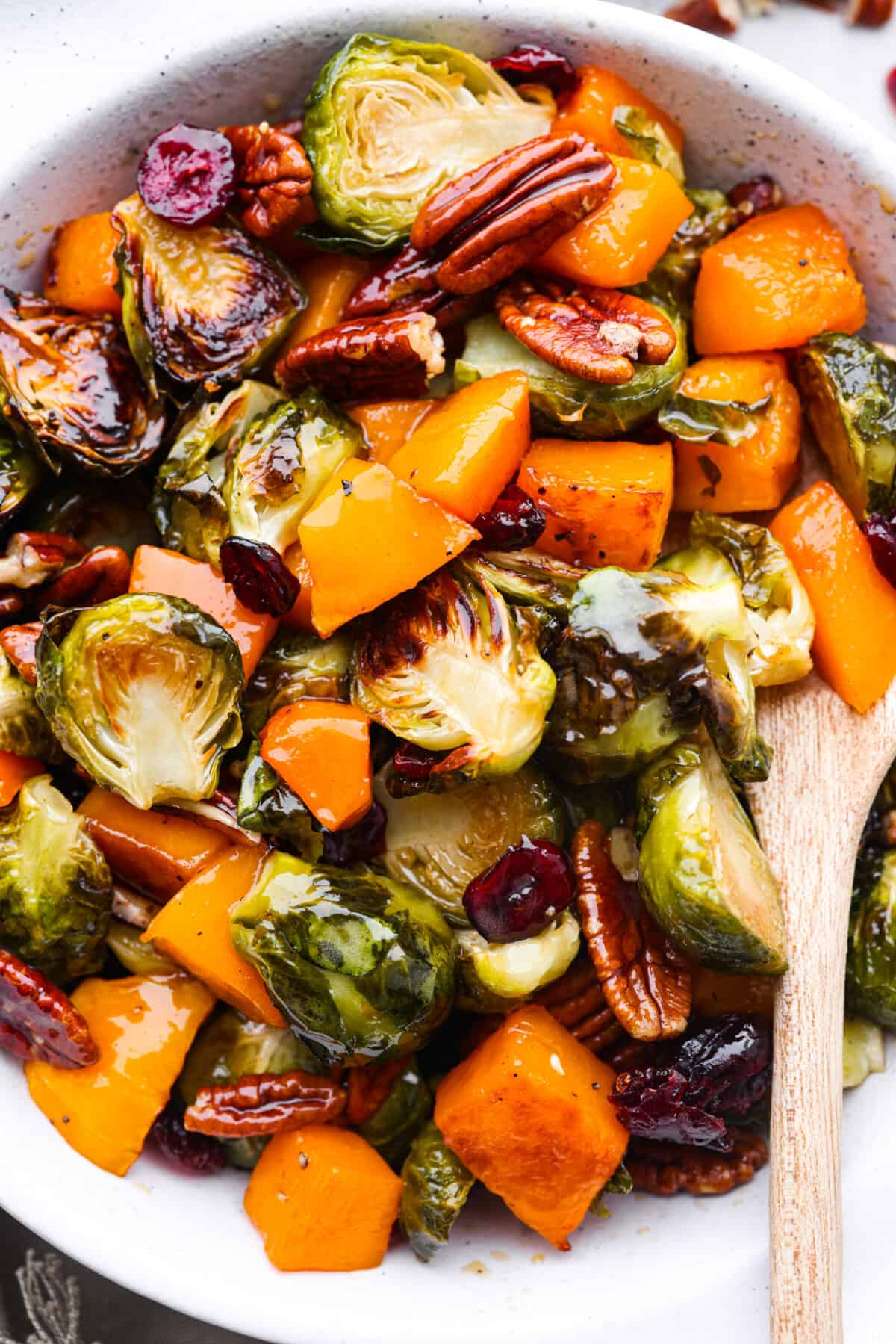 Simple to make and full of incredible flavor, maple roasted brussels sprouts and butternut squash is the perfect side dish. The veggies are crisp on the edges but soft and tender on the inside, tossed in a delicious maple glaze and surrounded by crunchy pecans and cranberries. Closeup of maple roasted vegetables in a white baking dish.